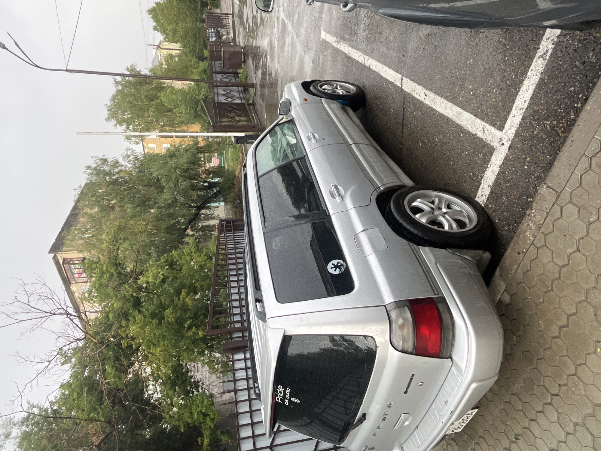 The image shows a silver Subaru Forester parked on a wet, paved surface, likely a parking lot in the city of Oskemen, Kazakhstan. The sky is overcast, and it appears to have been raining recently, as evidenced by the water on the ground and the car. The car has a spoiler on the rear hatch and several stickers on the rear window, including one that reads "Pride Car audio." A license plate with Cyrillic characters is visible on the rear bumper. In the background, there is a metal fence, trees, and apartment buildings, consistent with an urban environment. The lighting is dim due to the cloudy weather.