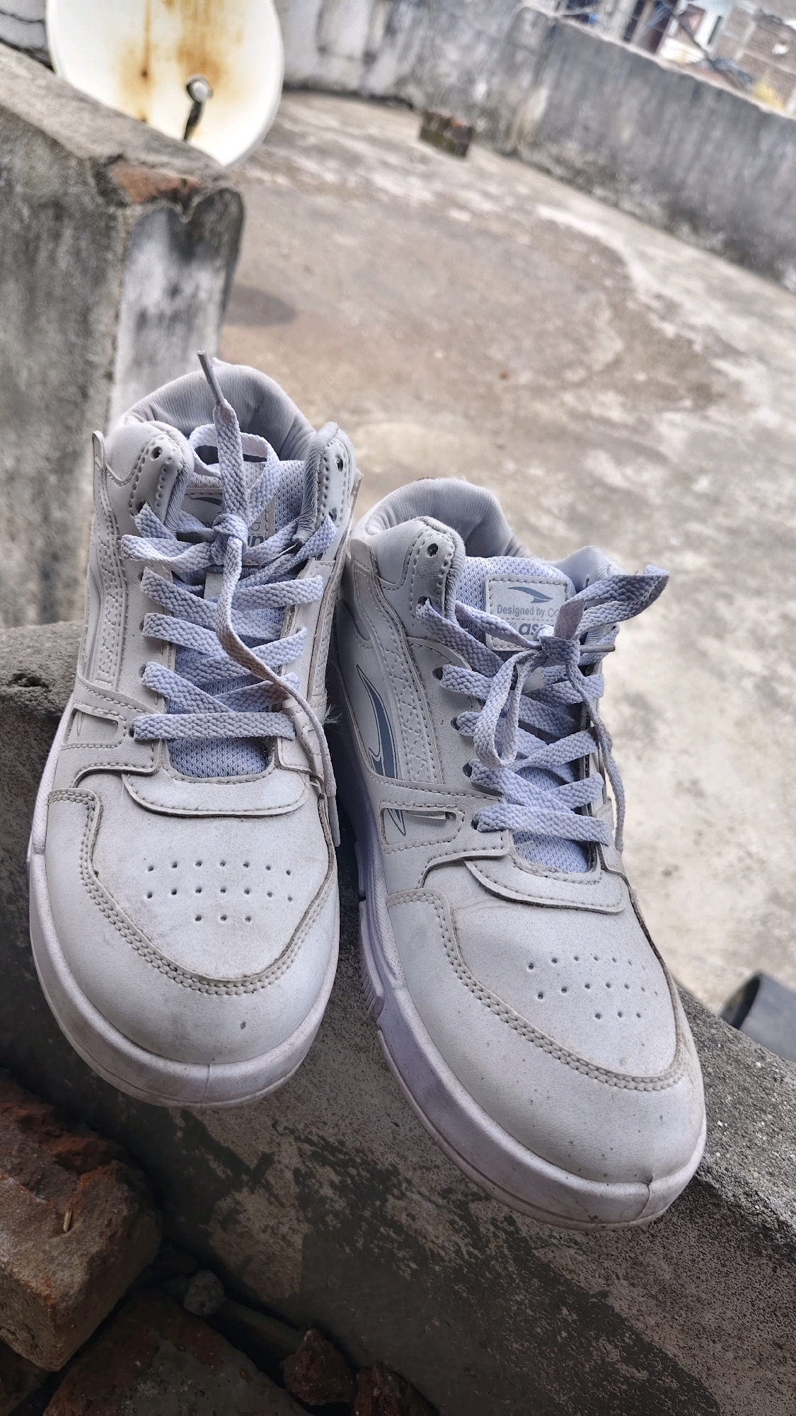 A pair of white, high-top sneakers with light purple laces are placed on a concrete ledge outdoors. The sneakers appear to be worn and are slightly dusty. The right sneaker has a logo that reads "Designed by C. AS." The background shows a rooftop with a satellite dish and various concrete structures, suggesting an urban or semi-urban environment. The ground is a textured concrete surface. The scene is lit by natural daylight, casting subtle shadows. The overall impression is one of a casual outdoor setting, possibly a rooftop in Thikri, India, where the shoes are placed for airing or storage.