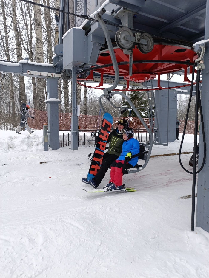 This image depicts a winter scene at a ski resort in Svetloe, Russia. Two individuals are seated on a grey metal two-person chairlift, ascending a snow-covered slope. The individual on the left, an adult, wears a dark green and black jacket, dark pants, and a helmet with goggles. They are holding a blue and black snowboard vertically, displaying "CAPITA" in orange lettering. The individual on the right, a child, wears a bright blue jacket, red ski pants, a helmet with goggles, and has yellow-green skis attached to their feet, suspended just above the snow. The ski lift's overhead mechanism features a large red bullwheel assembly supported by grey metal structures and pulleys. In the background, snow-dusted deciduous trees with light bark and some evergreens are visible. Another person carrying a snowboard is walking on the snowy terrain further up the slope. The ground is covered in white snow, showing tracks from previous activity near the lift's loading area.