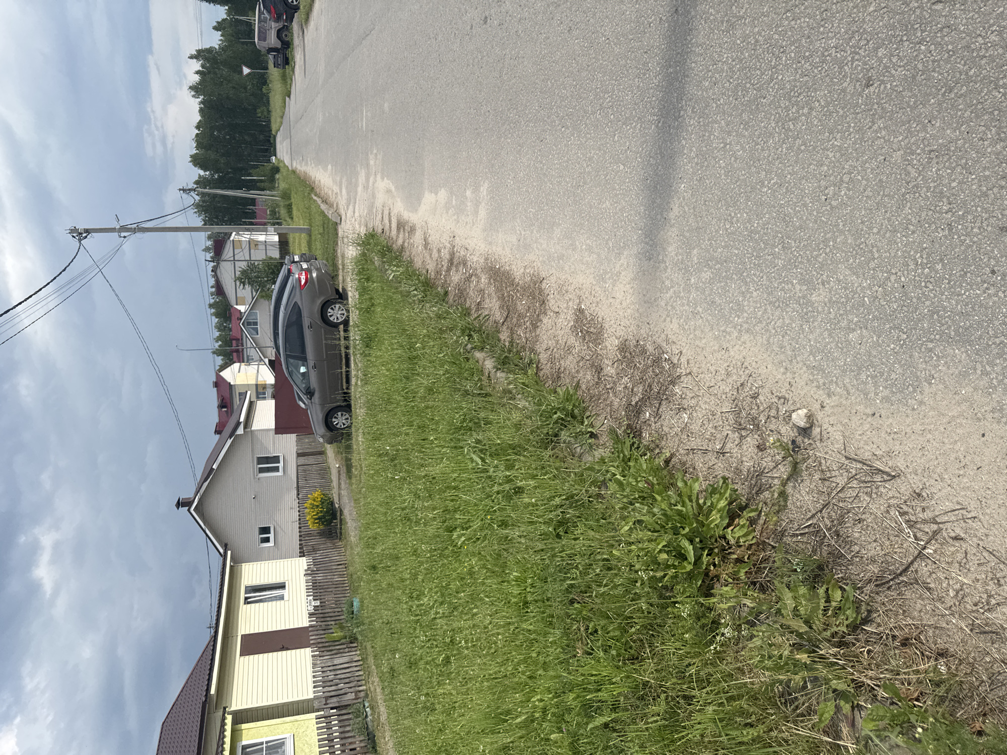 The image depicts a street scene in the village of Krasnye Baki, Russia, under a cloudy sky. The foreground features a gravelly verge with tall, green grass and some dry vegetation. A paved road runs diagonally from the lower right to the upper center of the frame. On the left side of the road, a row of houses with traditional wooden fences is visible. The houses have light-colored siding and dark roofs. A gray sedan is parked in front of one of the houses, next to a wooden fence. Further down the road, more houses and trees are visible, and power lines stretch across the scene. Several other vehicles, including a dark SUV and a lighter-colored utility vehicle, are parked on the right side of the road in the distance. The overall impression is one of a quiet, rural residential area.