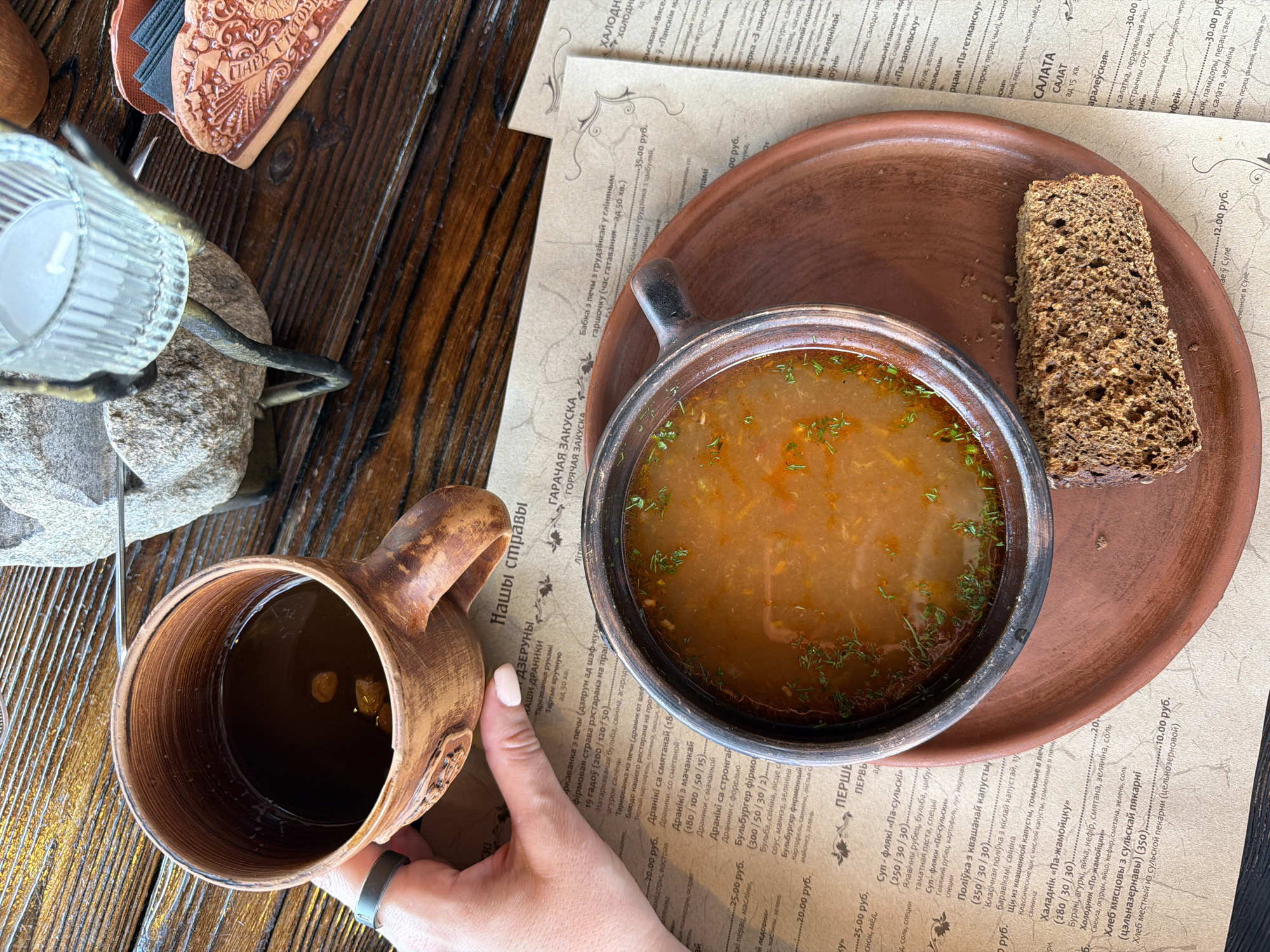 The image depicts a rustic, wooden table setting in what appears to be a traditional Belarusian restaurant in Літвенскі сельскі Савет, Belarus. In the foreground, a brown, earthenware bowl of hearty soup, garnished with fresh herbs, sits on a matching plate. A slice of dark, dense bread rests on the side of the plate. To the left, a hand with a silver ring is holding a brown, earthenware mug filled with a dark beverage, possibly tea or coffee, with some dried fruit floating in it. In the background, a decorative stone candle holder with a glass votive is visible, along with a napkin holder made of decorative clay. The table itself is made of dark, distressed wood, contributing to the overall rustic ambiance. The menu, with Cyrillic script, is partially visible beneath the bowl and mug.