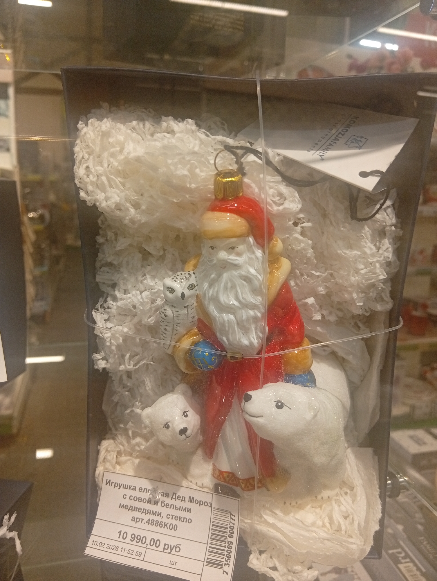 An indoor, eye-level, medium shot captures a glass Christmas ornament featuring Ded Moroz (Grandfather Frost) with a white owl on his shoulder and two polar bears at his feet. The ornament is housed in a dark box, filled with white shredded paper for cushioning. A white tag with Russian text and a barcode is attached to the box. The tag reads "Игрушка елочная Дед Мороз с совой и белыми медведями, стекло арт.4886К00 10 990,00 руб 10.02.2026 11:52:59 шт". The setting appears to be a retail store. The overall lighting is bright and diffused, typical of an indoor retail environment. The image is focused on the ornament and its packaging, with a blurred background of store merchandise. There are no people visible in the frame.