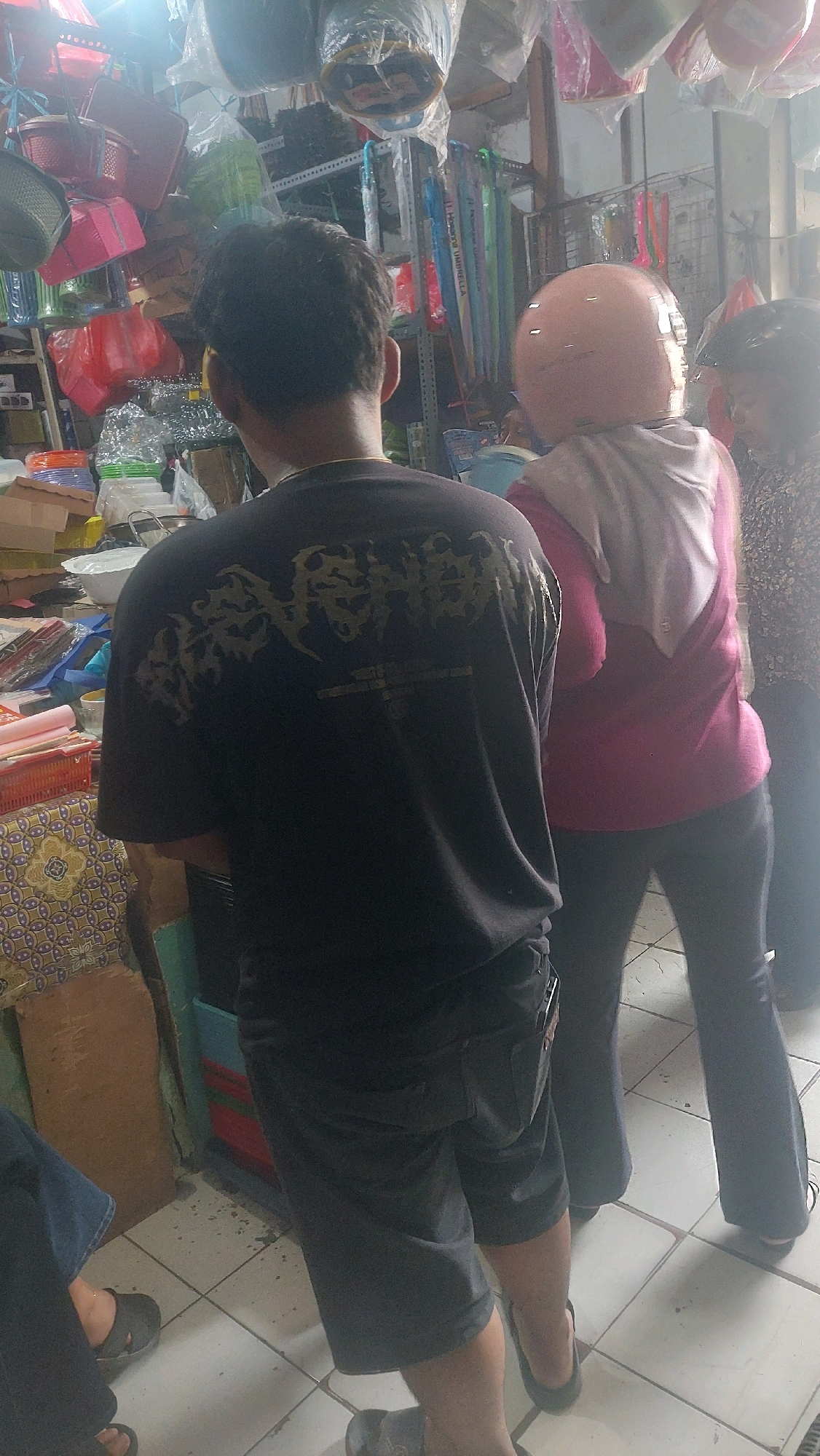 This is a medium-shot, full-color, interior photograph taken at an angle of three individuals in what appears to be a shop or market stall in Madiun, Indonesia. The background is cluttered with various items, including plastic containers, baskets, and merchandise hanging from the ceiling, suggesting a retail environment. The lighting is bright, with a strong light source visible at the top left of the image, possibly from the sun or an overhead lamp, creating some glare and overexposure in that area.
In the foreground and slightly to the left is a person from the back, wearing a black t-shirt with a gold, ornate graphic design. They have dark, short hair. This individual is wearing dark shorts and appears to be standing.
To the right of the first individual, and slightly behind them, is another person wearing a pink t-shirt and black pants. They are also facing away from the camera and are wearing a light pink helmet. A light-colored scarf or hijab is draped over their shoulders.
To the far right, a third person is visible from the side, wearing a patterned blouse and dark pants. They are looking towards the second individual.
The floor is tiled with light-colored square tiles, and there are some dark smudges on the floor. The overall impression is of a busy marketplace or a small retail establishment in Madiun, Indonesia.