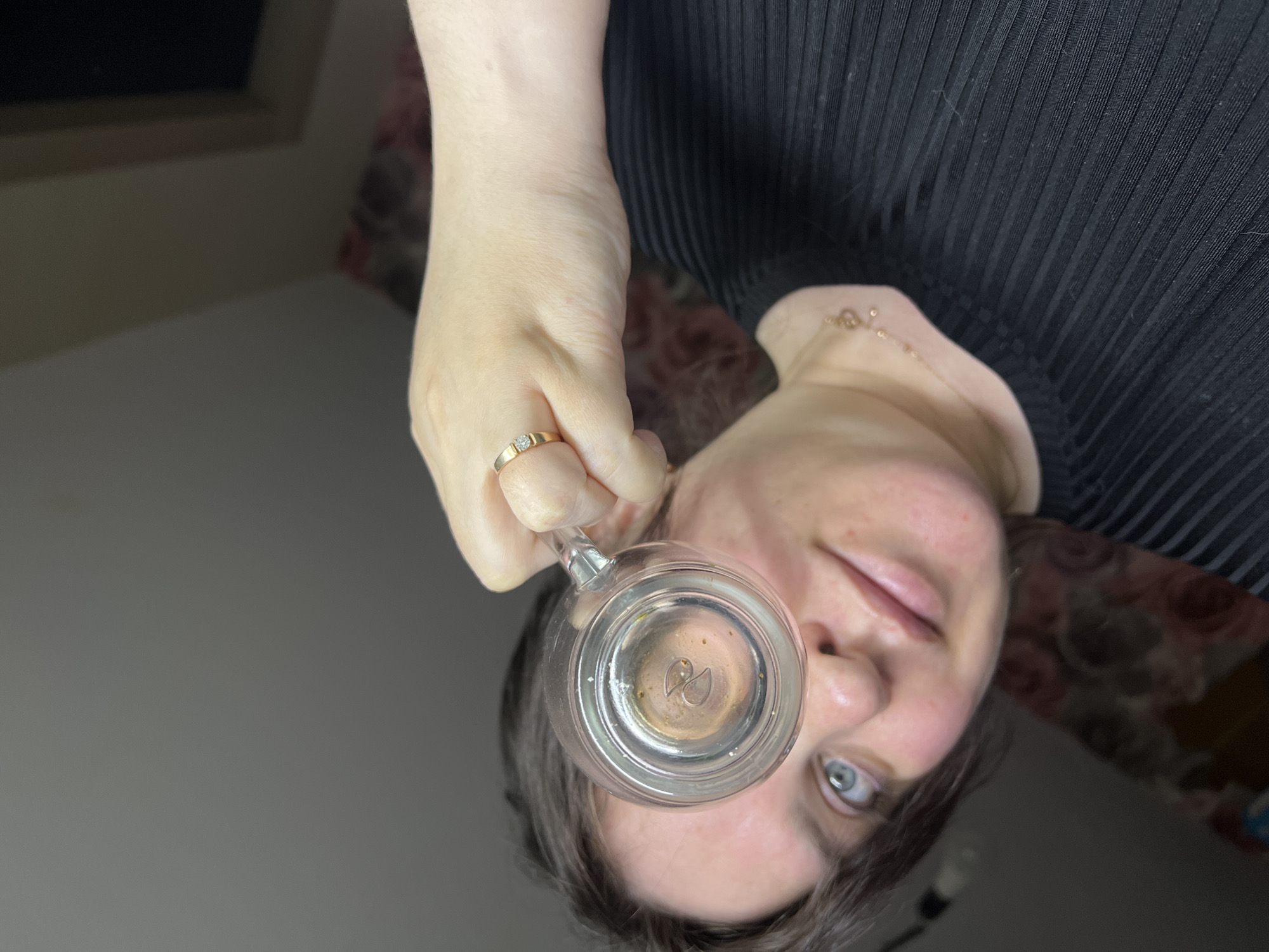 A person is holding a glass mug filled with a small amount of liquid in front of their face. The person has blue eyes and dark hair. They are wearing a black ribbed top and a gold ring with a diamond on their finger. A gold chain necklace is visible around the person's neck. The background includes a floral pattern and a gray surface. There is no discernible information to suggest the image was taken in Noviy, Russia.
