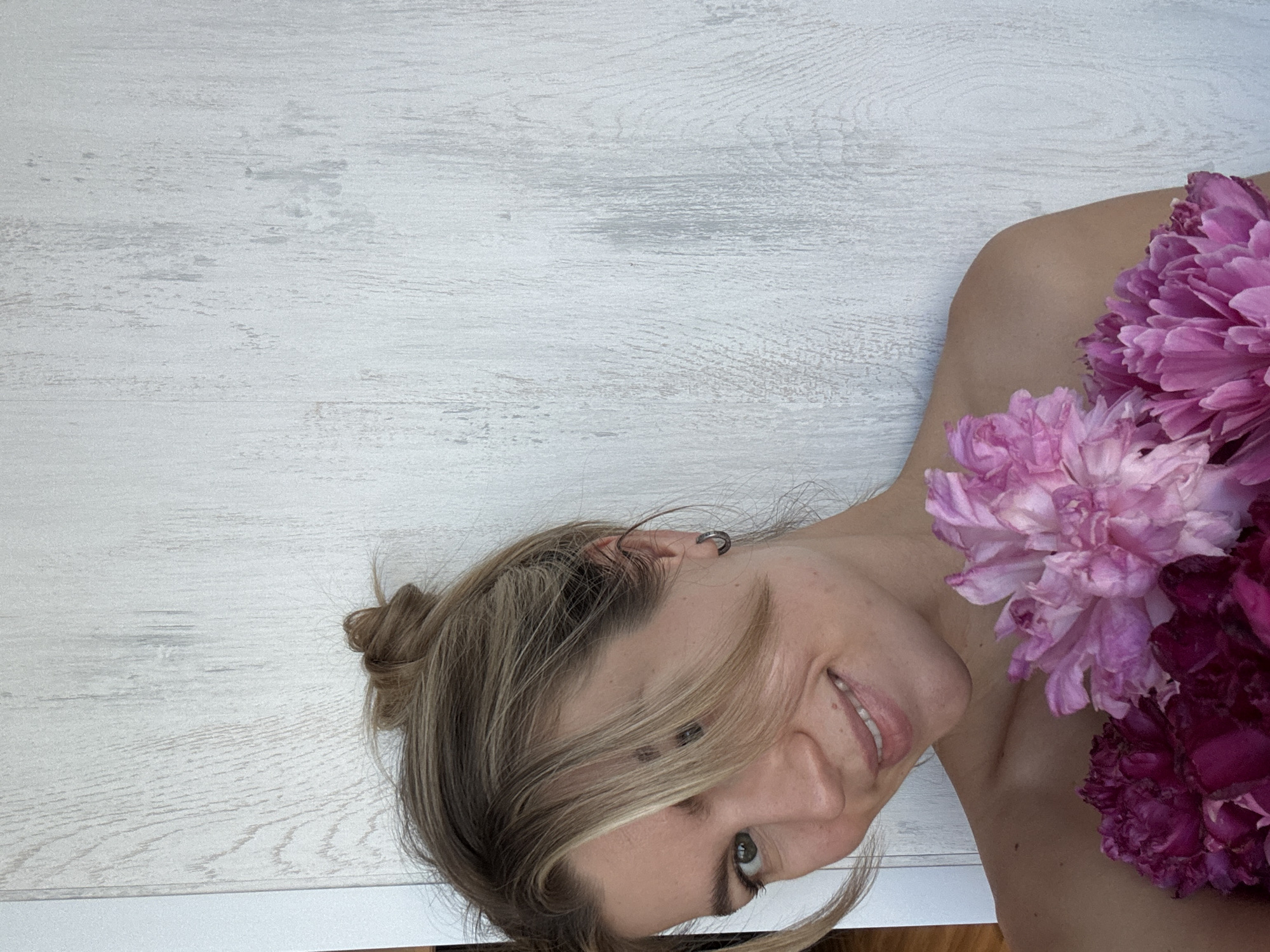 A fair-skinned woman with blonde hair pulled into a messy bun smiles at the camera. Her left eye is partially covered by bangs, and she is wearing a small hoop earring in her left ear. She holds several pink and magenta peonies in front of her bare shoulders. The background is a light wood-paneled wall. The scene is indoors, with no indication of its specific location within a city.