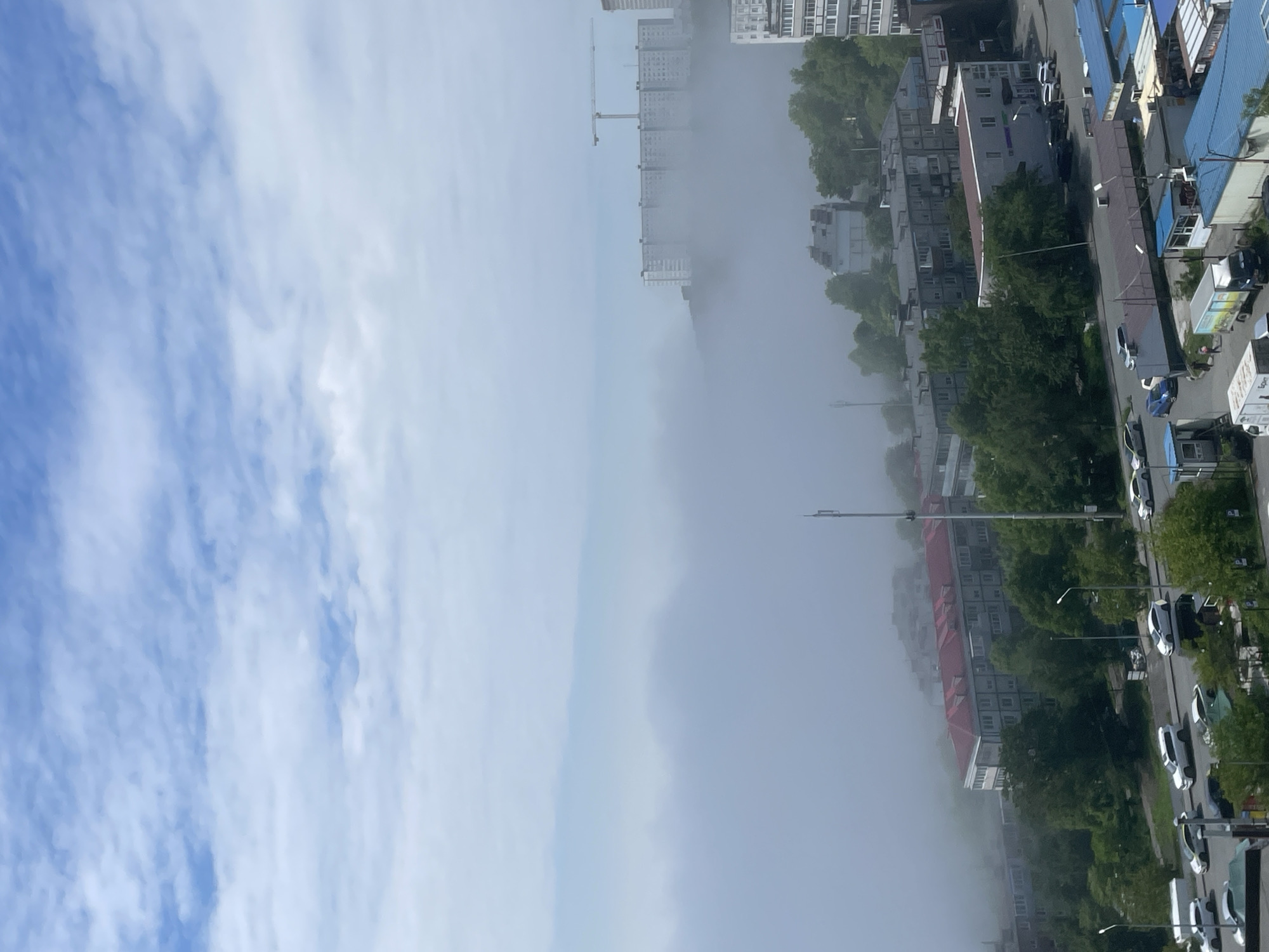 The image depicts a foggy morning in Vladivostok, Russia. The scene is dominated by a thick layer of fog that obscures the lower parts of buildings and trees. In the foreground, a street lined with parked cars and some kiosks is visible. Further back, apartment buildings of varying heights are partially shrouded in fog, with a construction crane visible above one of the larger buildings on a hill. The sky above is a mix of blue and white clouds, indicating the fog is a low-lying phenomenon. The overall impression is one of a misty, urban environment.
