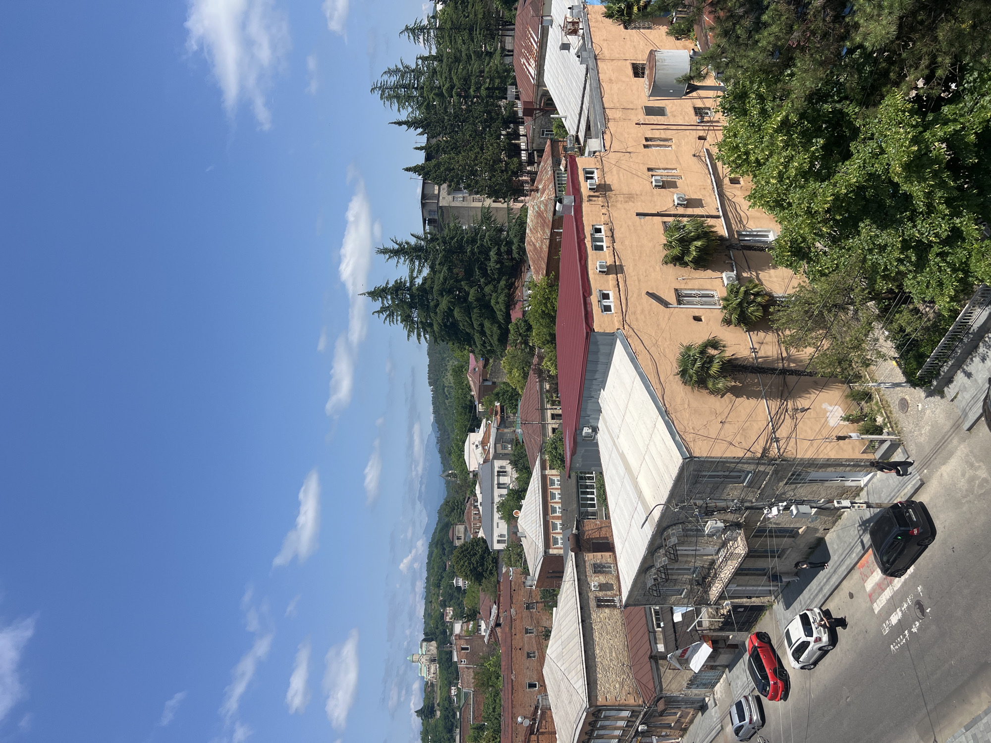 The image shows a street scene in Kutaisi, Georgia, on a sunny day with a blue sky and scattered white clouds. The view is from an elevated perspective, looking down onto a street lined with buildings and trees.
In the foreground, a street runs horizontally across the bottom of the image. Several cars are parked and driving on the street. A black car is parked on the right side of the street, while a white SUV and a red hatchback are parked further up on the left. A silver sedan is parked in the distance on the left. Pedestrians are visible on the sidewalks.
The buildings lining the street are of varying architectural styles and ages. Some are made of brick, while others are stuccoed and painted in warm tones like orange and yellow. Many buildings have multiple stories and feature windows, balconies, and air conditioning units. Several trees, including palm trees and mature evergreen trees, are interspersed among the buildings, adding greenery to the urban landscape.
In the background, more buildings and trees are visible, extending towards rolling hills covered in lush vegetation. In the far distance, mountains can be seen under the hazy sky. A prominent building with a dome and a green roof, likely a church or cathedral, sits atop a hill in the upper left portion of the image, overlooking the city. The overall impression is a vibrant and lived-in urban environment with a blend of traditional and modern elements, set against a backdrop of natural beauty.