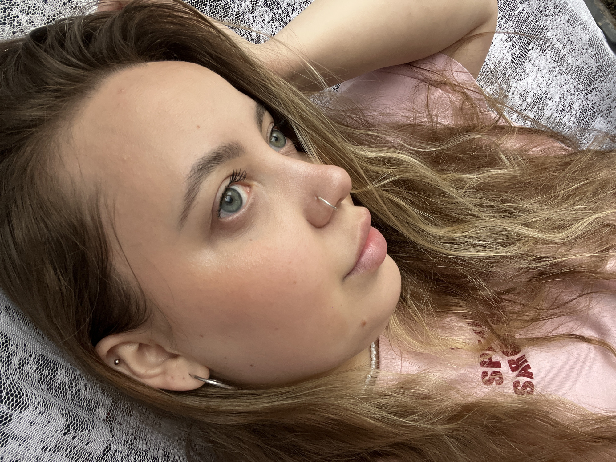 A young woman with light brown and blonde hair lies on a white crocheted blanket or tablecloth. She is wearing a pink t-shirt and a pearl necklace. She has a silver nose ring and a silver hoop earring, as well as a smaller stud earring in her ear. The location is not discernible from the image.