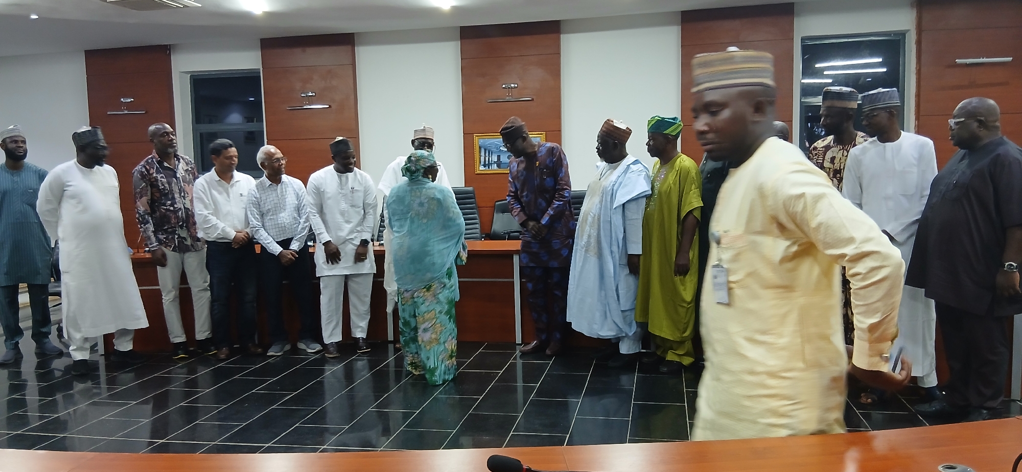 In the city of Unguwan Sanusi, Nigeria, a group of twelve individuals, predominantly men, are gathered indoors in what appears to be a meeting room or conference hall. The room features dark wooden paneling on the walls and a tiled floor with a reflective surface. The lighting appears to be artificial, emanating from overhead fixtures.
The individuals are standing in a semi-circle, facing towards the center of the room. Their attire varies, with some wearing traditional Nigerian clothing such as agbadas and caps, while others are dressed in shirts and trousers. Several individuals are wearing distinct headwear, including caps and turbans. One woman, in the center of the group, is wearing a light blue and green patterned headscarf and dress.
The people are in various poses: some are standing erect and looking forward, others are engaged in conversation, and one individual in the foreground is in motion, partially obscuring the view of others. There are office chairs visible behind some of the individuals, and a large wooden table is in the foreground, with a microphone on it. The overall atmosphere suggests a formal gathering or meeting.