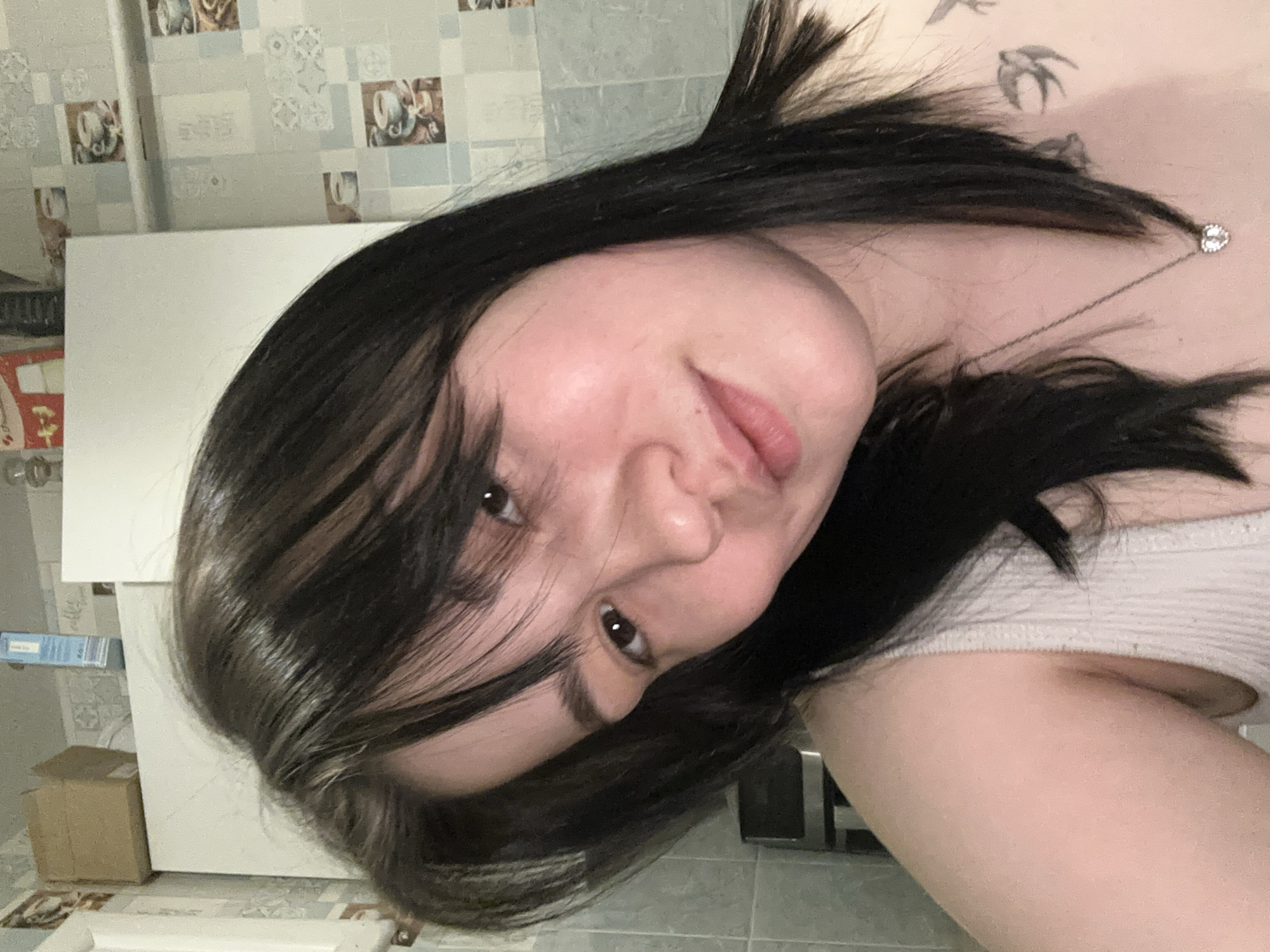 A close-up, rotated portrait captures a young woman looking directly at the camera, taken indoors in Chaikovsky, Russia. Her head is tilted, giving the impression she might be lying down or leaning back. She has long, dark hair, with some lighter strands or highlights, partially obscuring her forehead and left eye. Her skin is fair, and her lips are slightly parted.
She wears a light-colored, possibly white or off-white, ribbed tank top or camisole. A delicate silver chain necklace with a small, sparkling heart-shaped pendant hangs on her chest. A tattoo depicting two birds in flight is visible on her upper chest/shoulder.
The background features a wall adorned with tiles or wallpaper patterned with light blue/grey squares alternating with squares containing images of coffee cups and other decorative elements. To the left of her head, a large white rectangular object, potentially a canvas or a piece of furniture, is visible. Below this, a small cardboard box is faintly discernable. A thin, vertical pipe or rod is also present in the upper left background. The lighting is soft and even, highlighting her face. No text is visible within the image. The specific time of day or weather conditions cannot be determined.