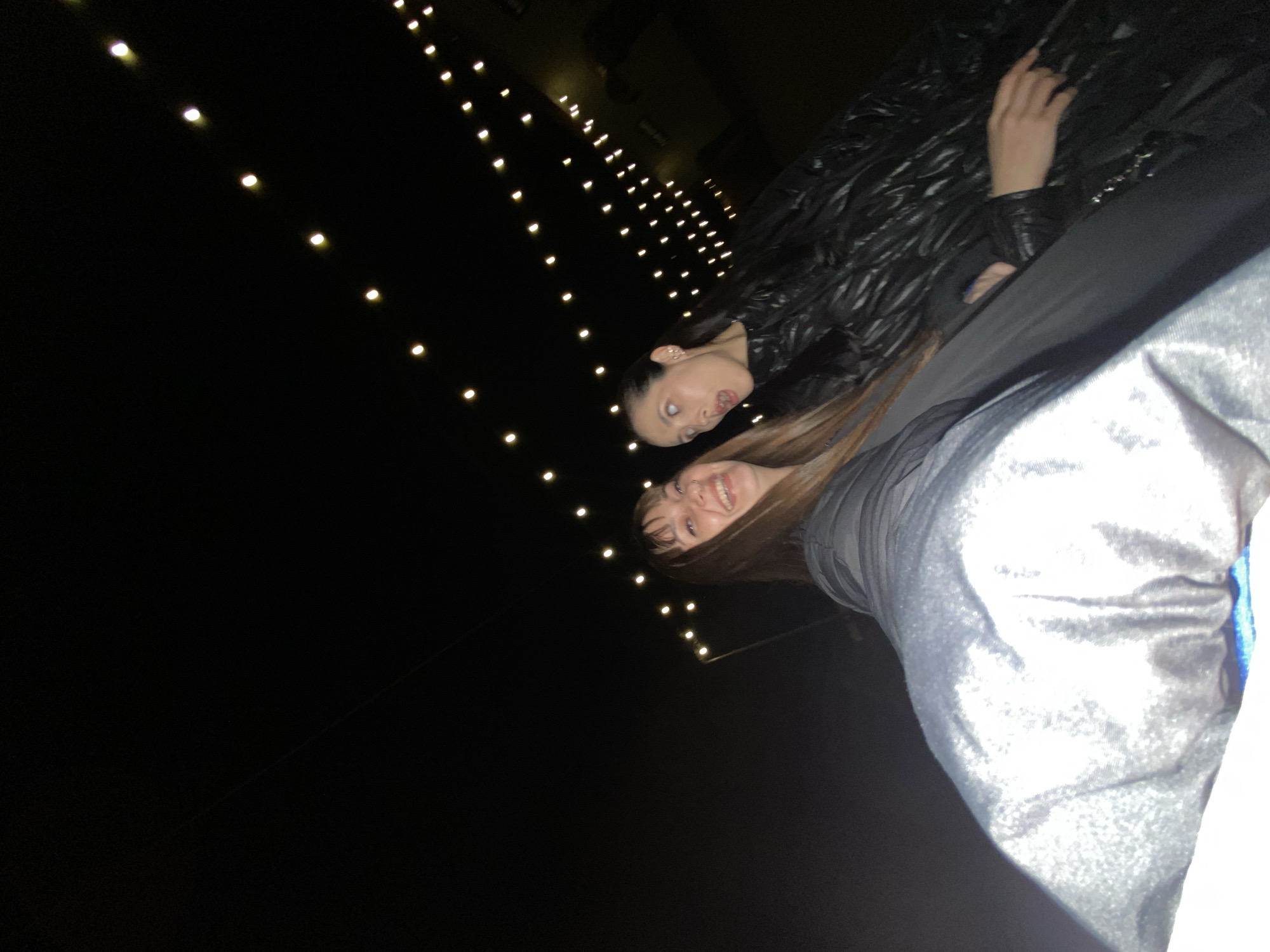 The image shows two young women posing for a photo in an indoor or covered setting at night in Votkinsk, Russia. The background is predominantly dark, illuminated by numerous small, warm-toned lights arranged in diagonal lines, suggesting a decorative ambiance like a restaurant, bar, or event space.
The woman closer to the camera, positioned on the right side of the frame, has long, straight brown hair and is smiling broadly. Only her head, upper body, and a portion of her arm are visible. She is wearing clothing made of a grey or silver fabric with a subtle sheen, possibly velvet.
Behind her and slightly to the left is the second woman, with dark hair, looking towards the camera. She is making a playful expression, with her eyes rolled upwards and her tongue slightly extended. She is dressed in a dark, possibly black, puffy or quilted jacket. Her hands are also visible, one resting at her side and the other slightly raised.
The photograph is taken from a low, tilted angle, giving it a casual and candid feel. The visible emotions are cheerful and playful. The dark surroundings confirm that the image was taken at night. No text is visible.