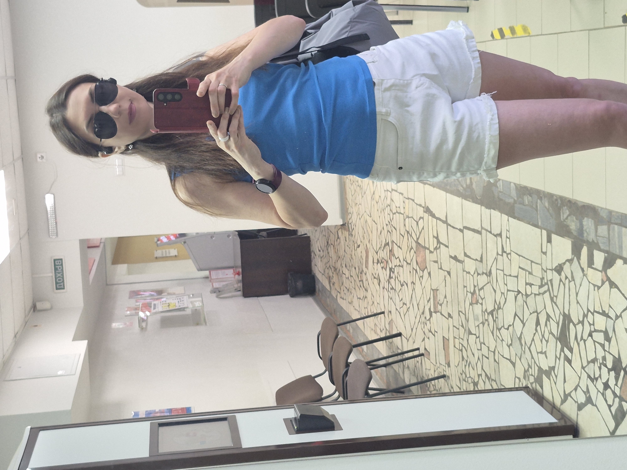 The image depicts a woman taking a mirror selfie in what appears to be an indoor public space, possibly a transportation hub or administrative building. The woman is wearing dark sunglasses, a blue top, and white shorts. She is holding a dark red smartphone, presumably to take the photo, and is wearing a smartwatch on her left wrist. Behind her is a grey backpack.
The background reveals a tiled floor and a wall decorated with mosaic patterns. There are several chairs lined up near the wall. A desk-like structure with a dark base is visible in the foreground. On the left of the frame, there is a wall with signage in Russian, possibly indicating an exit ("ВЫХОД"). The room seems to be well-lit and appears to be in a location with an older architectural style.