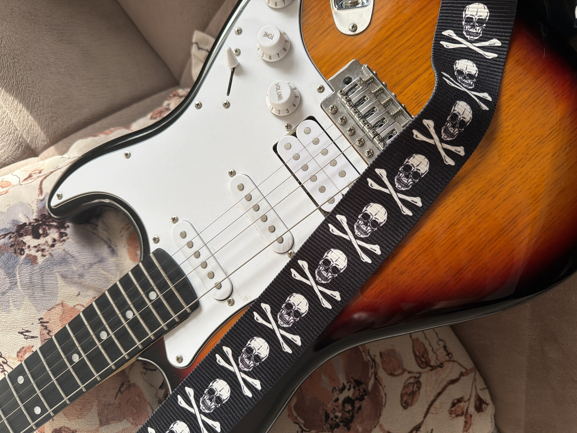 This media file is a close-up, overhead shot of an electric guitar, likely a Fender Stratocaster style, resting on a soft surface that appears to be a couch or a large cushion with a floral pattern. The guitar has a sunburst finish, transitioning from a deep red to a warm orange and yellow. Its pickguard is white, and it features three single-coil pickups.
A black guitar strap, adorned with a repeating pattern of white skulls and crossbones, is draped diagonally across the body of the guitar, obscuring some of the pickups and the bridge. The strap itself is a notable detail, suggesting a particular aesthetic or musical preference. Visible controls on the guitar include two white knobs labeled "TONE" and "VOLUME," each with numerical markings from 0 to 10. The neck of the guitar is visible, with its dark fretboard and silver frets.
The lighting suggests it might be daytime, with soft shadows cast by the guitar, particularly on the strap. There are no people present, and the focus is solely on the musical instrument and its accessories. The setting appears to be indoors, within a domestic environment. No text other than the labels on the control knobs is visible. The overall impression is one of a stylish and well-maintained musical instrument.