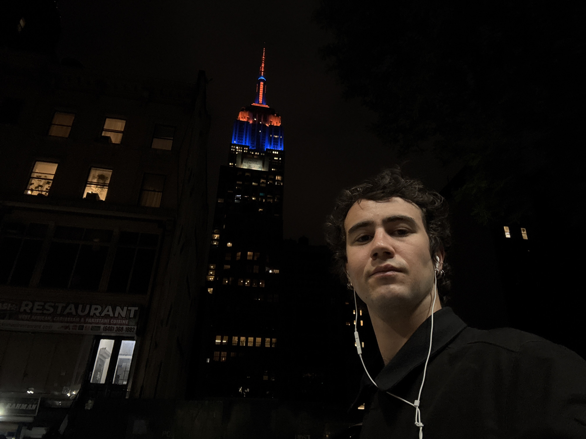 A nighttime medium shot shows a man wearing earphones on a city street in New York City. In the background is the Empire State Building, lit with blue and orange lights, suggesting a possible event or celebration. The man is wearing a dark shirt and faces forward, and his expression is neutral. To the left of the frame is the side of a building with illuminated windows and a restaurant sign that reads "RESTAURANT." The scene is dark, with the city lights providing the only illumination.