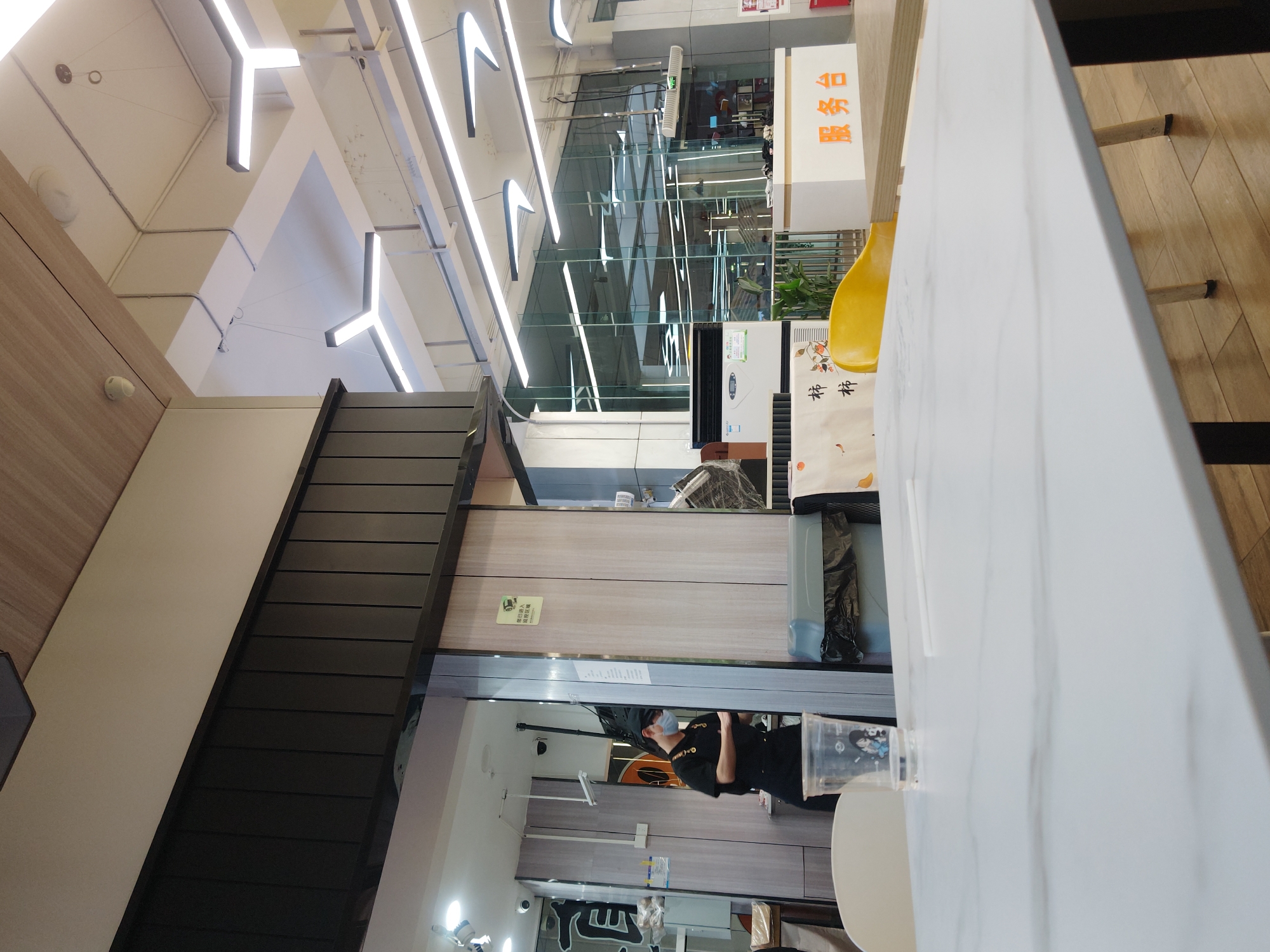 The image presents an interior view of a modern commercial space, possibly a cafe, restaurant, or a section of a food court, located in Chongqing High-tech Zone, China. The environment is well-lit with cool, artificial light emanating from distinctive geometric LED fixtures, shaped like angular 'L's, mounted on the ceiling alongside exposed conduits.
To the left, a wall features light wood paneling above a lighter section, with a prominent dark, slatted structure, possibly a decorative wall element or part of a staircase, descending into the lower left of the frame.
The central and right portions of the image are defined by a large glass partition. Beyond this glass, another interior space is visible, featuring light wood-paneled walls and various equipment. A person, wearing a black uniform and a surgical mask, stands within this area, visible through the glass. A small green sign on the wall behind them reads "请勿倚靠 谨防滑跌" (Please do not lean, beware of slipping).
In the foreground on the right, a white table with a subtle gray marble pattern is prominently featured. A bright yellow chair is partially visible behind the table. Next to the chair and behind the table, there's a white appliance or display stand, potentially an air purifier or cooler, with a poster attached bearing two Chinese characters, "柿" (Shì), meaning "persimmon." Further in the background, a white stand displays the Chinese characters "谷茶屋" (Gǔ Chá Wū), which translates to "Valley Tea House" or "Grain Tea House," likely indicating the name of the establishment. A clear plastic cup with a printed image is also visible near the table's edge.
The scene depicts a clean, contemporary setting typical of a modern commercial establishment. There is no specific activity taking place beyond the presence of a staff member. The bright indoor lighting does not suggest a particular time of day.