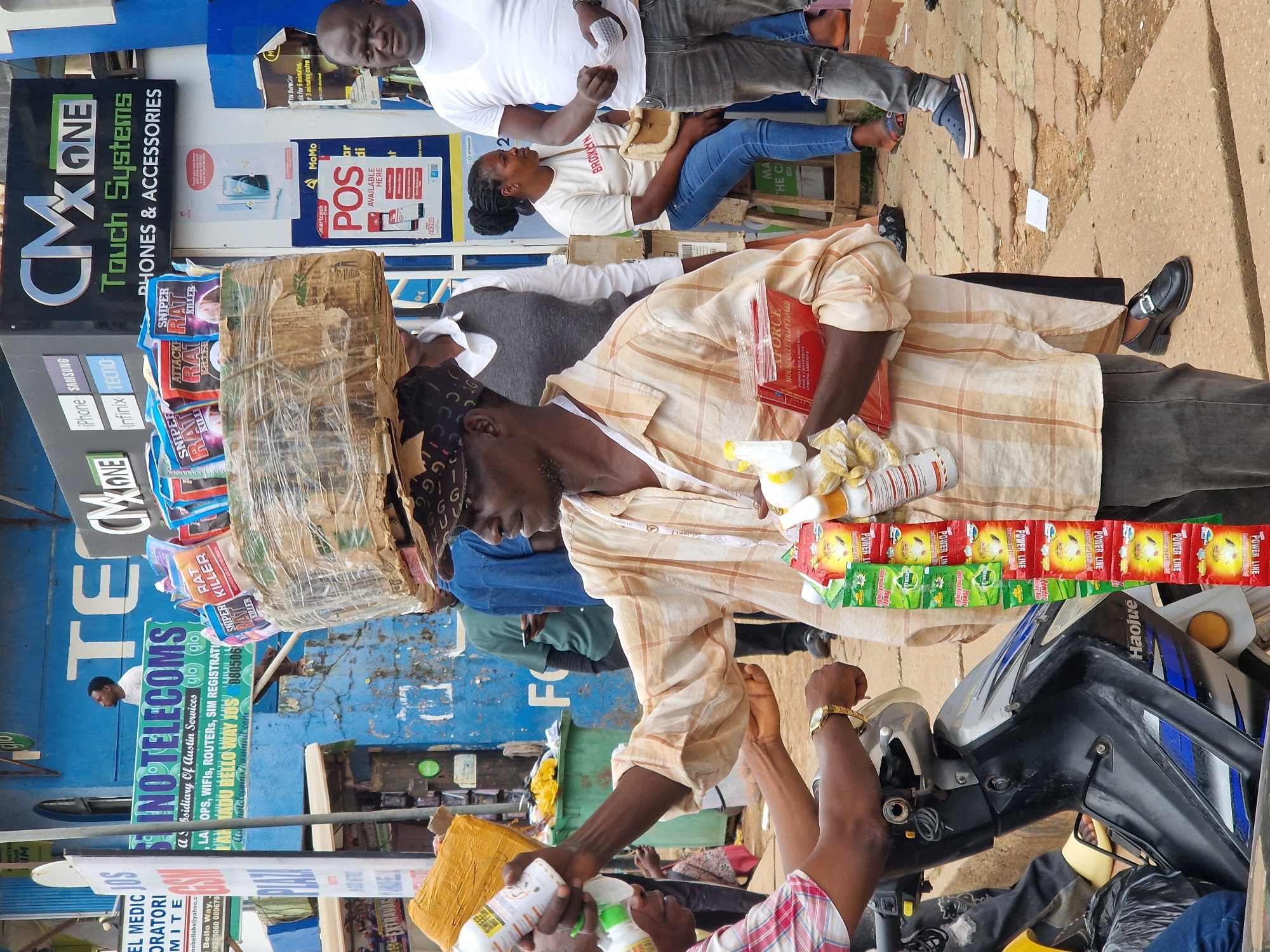 This is a photograph of a busy street scene, likely in a market or commercial area, with an emphasis on the sale of mobile phones and telecommunication services. The image is vertically oriented, giving a sense of looking up at the scene.
In the foreground, a man is being handed what appears to be a bottle of insecticide or cleaning spray. He is wearing a light-colored, striped, short-sleeved shirt and a dark head covering. He is holding a large, cylindrical package wrapped in plastic and cardboard on his head. Beside him, another person, partially visible, is wearing a plaid shirt and also holds a bottle.
Behind them, a variety of stalls and signs are visible. One prominent sign, in black and green, advertises "CMX ONE Touch Systems PHONES & ACCESSORIES" and lists various phone brands like "iPhone SAMSUNG Infinix TECNO." Another blue sign reads "INO TELECOMS" and mentions services like "OPS, WIFIS, ROUTERS, SIM REGISTRATION." A red poster is displayed with "MoMo POS AVAILABLE HERE."
Several people are present in the background, including a man in a white t-shirt and jeans standing near a sign, and a woman in a t-shirt and blue jeans sitting down. The ground is paved with cobblestones. The overall atmosphere appears to be that of a vibrant marketplace, with goods being displayed and transactions likely taking place. The time of day is not explicitly clear, but the lighting suggests it might be daytime. There are no obvious indications of the weather.