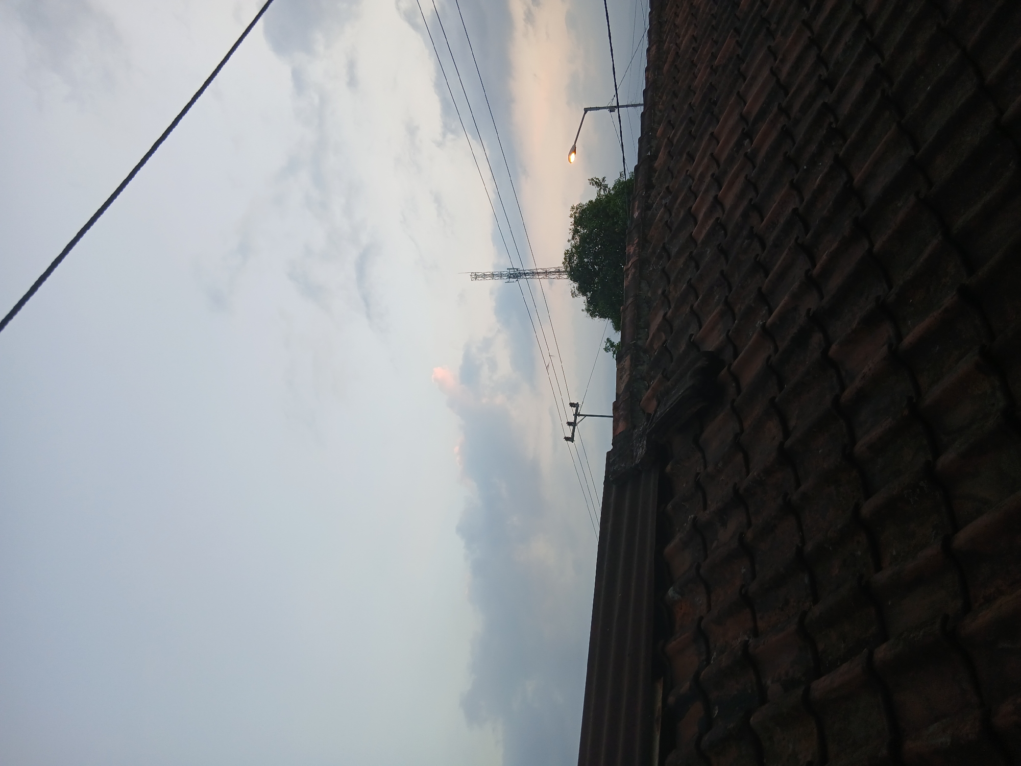 The image captures a rooftop perspective overlooking a scene in Kademangan Kulon, Indonesia, during what appears to be dusk or dawn. In the foreground, a dark, tiled roof dominates the lower half of the frame, showing rows of weathered, reddish-brown ceramic tiles. Some sections of the roof are corrugated metal, with a darker, possibly rusted, texture.
The middle ground features a cluster of trees, partially obscuring a tall, lattice-like communication tower in the distance. To the right of the tower, a streetlamp is illuminated, casting a warm glow. Overhead, multiple dark power lines crisscross the sky, supported by poles visible on the rooftops. The sky itself is a soft gradient of pale blue, with wispy clouds tinged with pink and orange, suggesting either sunrise or sunset. The overall atmosphere is tranquil and evokes a sense of a quiet residential area. No people or animals are visible in the frame.