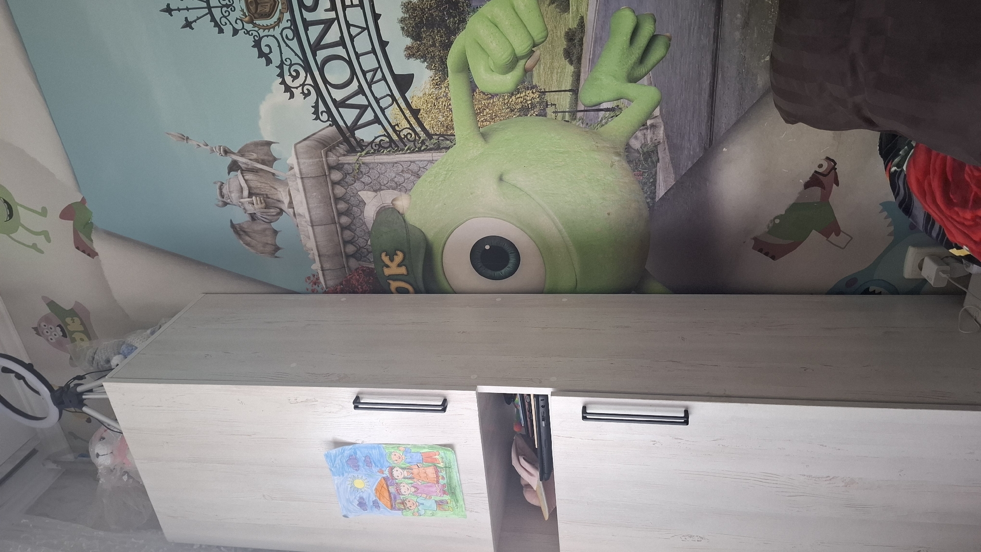 The image depicts the interior of a room, likely a child's bedroom, featuring a light-colored wardrobe with black handles. A child's drawing of a family is attached to the top door of the wardrobe. The lower section of the wardrobe has an open shelf containing a laptop and some pink fabric items. To the right of the wardrobe is a wall adorned with a large mural or wallpaper featuring characters from Disney/Pixar's Monsters, Inc. franchise. A prominent character visible is Mike Wazowski, the one-eyed green monster, partially obscured by the wardrobe. The mural also includes a gate with the inscription "MONS UNIV," suggesting Monsters University. The environment appears to be a domestic indoor setting. There is no discernible information in the image that would allow for the identification of its location within Shymkent, Kazakhstan.