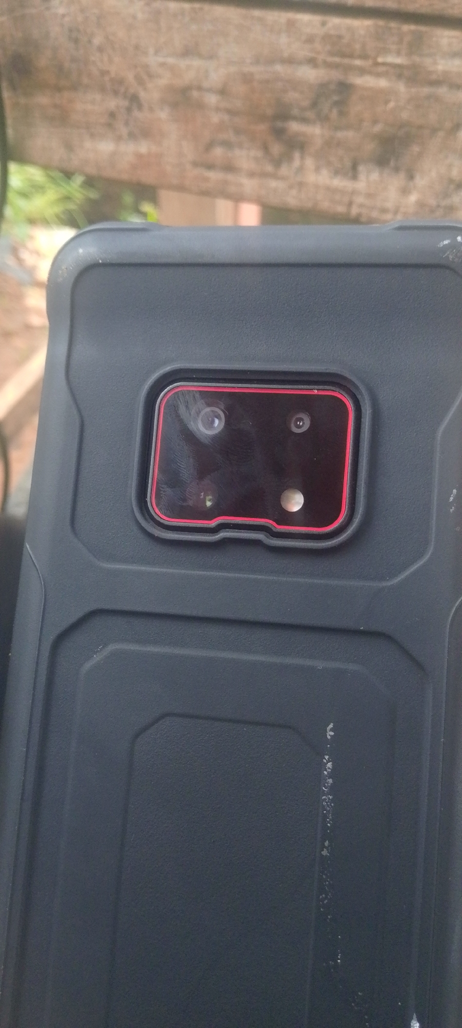 A close-up, overhead shot shows the rear camera module of a dark-colored smartphone case. The camera area is recessed within the case and features a rectangular housing with a red trim. Inside the housing are three visible lenses: two circular lenses in the top row, and one circular lens in the bottom row, which appears to be a flash or a depth sensor. The phone is lying on a weathered wooden surface, possibly part of a bench or railing, with blurred green foliage visible in the background. The overall lighting suggests an outdoor setting. There are no people or any specific markers indicating a location in Baissa, Nigeria.