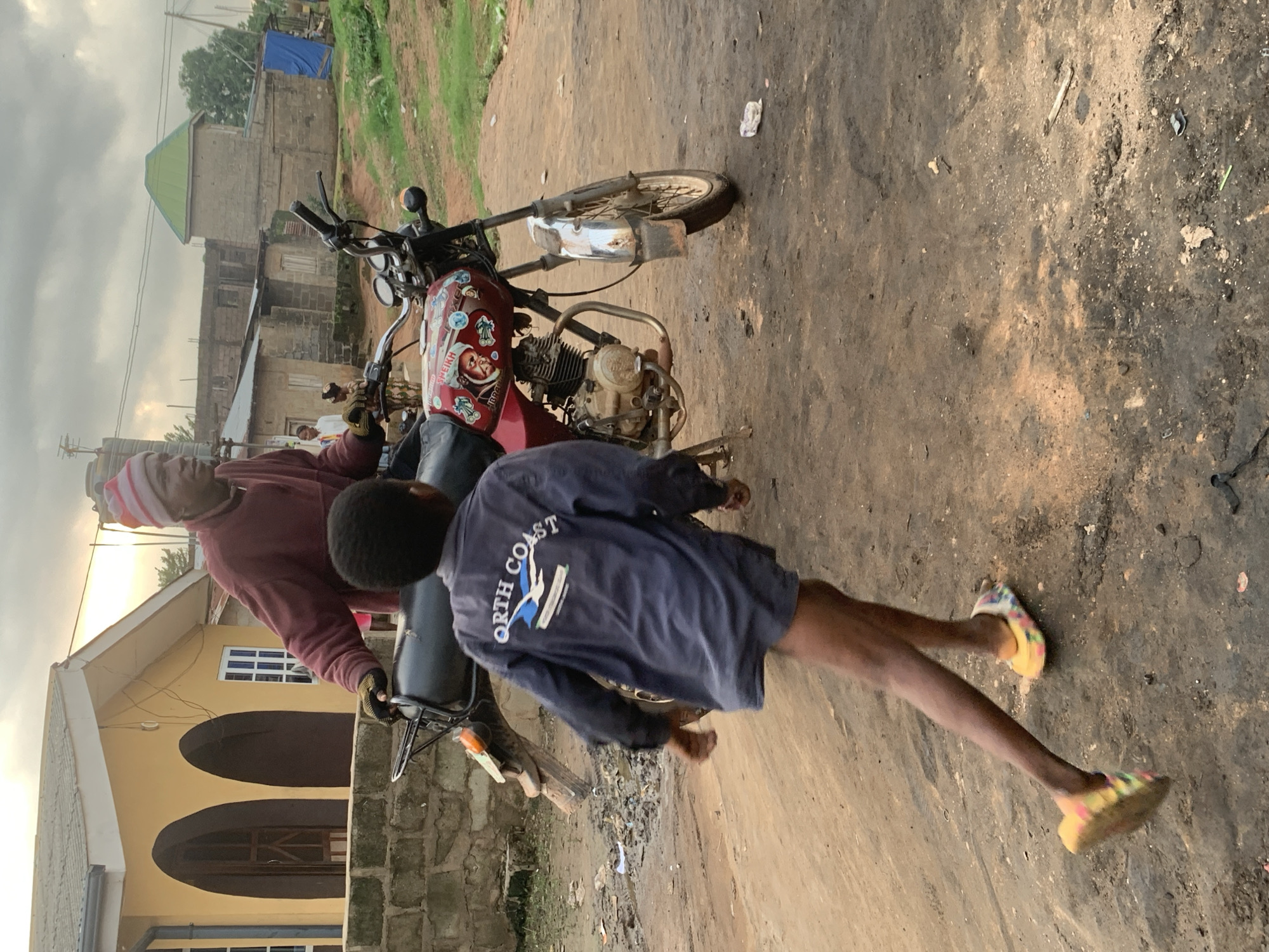 In Shagamu, Nigeria, the image captures a street scene with two individuals near a motorcycle. The person in the foreground, wearing a red maroon sweatshirt and a striped beanie, appears to be an adult. They are looking downwards and to the right. Behind them, a child, seen from the back, is wearing a dark blue sweatshirt with the text "NORTH COAST" and a bird graphic printed on it. The child is in motion, with their right leg extended, and is wearing colorful sandals.
The motorcycle is a prominent object in the scene, a red and silver model adorned with stickers. One sticker features a portrait of a man and text that includes "SHEIKH". The motorcycle is parked on a dirt-packed, uneven ground that shows signs of being a road or pathway.
In the background, a yellow building with arched doorways and a white-framed window is visible, along with other structures that suggest a residential or commercial area. The sky is overcast, indicating a cloudy or possibly approaching rainy weather, and the lighting suggests it could be late afternoon or early morning. The overall atmosphere appears casual, possibly depicting a moment of everyday life on a Nigerian street.