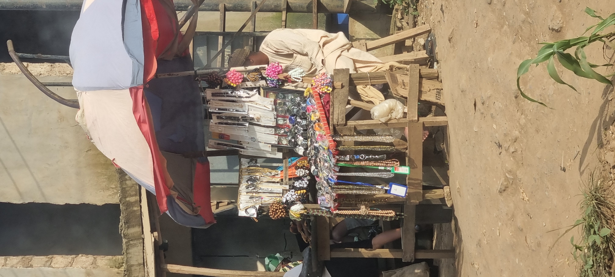 A street vendor sits behind a stall laden with watches and jewelry under a multi-colored umbrella. The stall, made of rough wooden planks, displays an assortment of watches, necklaces, bracelets, and hair accessories. The vendor, a man wearing a light-colored striped outfit and a cap, is seated on a wooden stool. The background shows a building with a rough, unfinished facade and scaffolding. The ground is dirt, with some sparse vegetation in the foreground. This scene is located on a street in Otta, Nigeria.