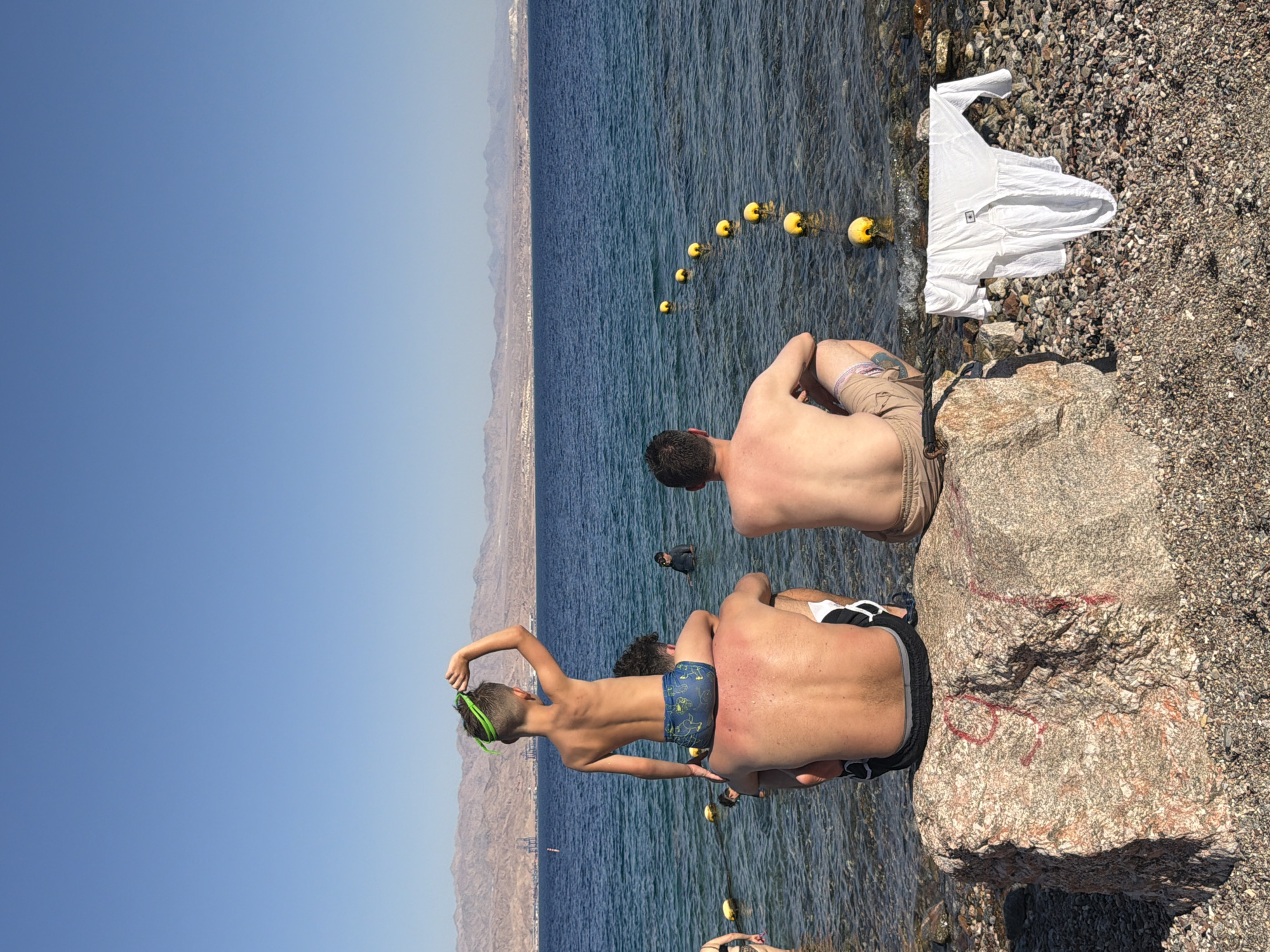 The image shows two people sitting on rocks at the edge of the sea, with a mountainous landscape in the background. One person, a young boy wearing green goggles and blue swim trunks, is sitting on the shoulders of another person. The second person, wearing brown shorts, is sitting on the rocks facing away from the camera. The water is calm and blue, with a line of yellow buoys extending into the distance. A white t-shirt is folded and placed on the rocky shore to the right of the frame. The sun is shining brightly, indicating it is daytime, likely warm weather given the attire and setting. The scene appears to be a recreational area, possibly a beach or a swimming spot. The presence of buoys suggests a designated swimming area or a marker for some activity in the water. The distant mountains and a hazy horizon suggest a coastal location. The overall mood appears relaxed and summery. There is no visible text in the image.