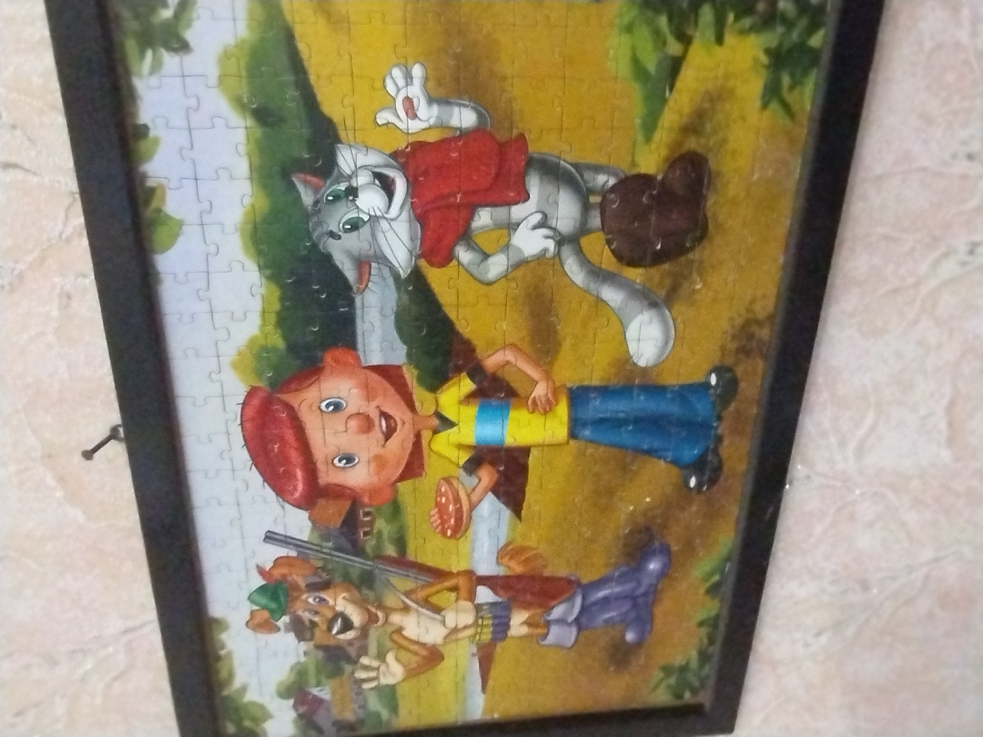 This is a framed jigsaw puzzle depicting a cartoon scene with three characters in an outdoor setting. On the left is a brown anthropomorphic dog wearing a green hat, a blue bandolier with bullets, a brown satchel, and large purple boots. The dog is holding a rifle and waving with its right hand. In the center is a cartoon child with red hair, wearing a yellow shirt with blue stripes and blue pants. The child is holding a circular, flat, reddish-brown object, possibly a cookie or pie, in their left hand. To the right is a grey anthropomorphic cat wearing a red scarf and brown boots. The cat is standing with its right hand on its hip and its left hand raised, also holding a small reddish object. The background of the puzzle shows a yellow-green grassy field with some trees and buildings in the distance, suggesting a rural or park-like environment. The puzzle is framed with a black border. The image is taken at an angle, showing the puzzle hanging on a wall with a textured, light-colored surface. There is no discernible location in the city of Samara, Ukraine.