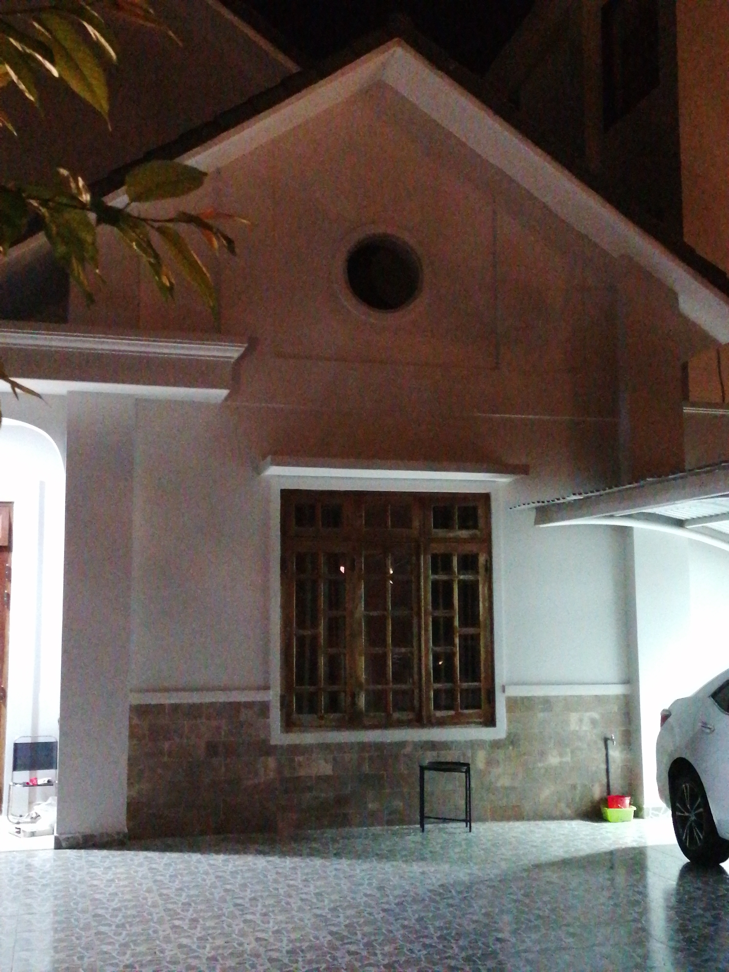 The image shows the exterior of a building at night, with a white car parked to the right. The building has a large wooden-framed window with multiple panes and a decorative stone facade below it. A small black stool is placed in front of the window. To the right of the stool, near the wall, are a red bucket and a green basin, with a black faucet and pipe above them. Leaves from a tree hang into the frame from the top left. The foreground is a tiled patio. The lighting suggests an outdoor setting at night, possibly a residential area in Da Lat, Vietnam, given the architectural style and the presence of the car and outdoor items. There are no people visible in the image.
