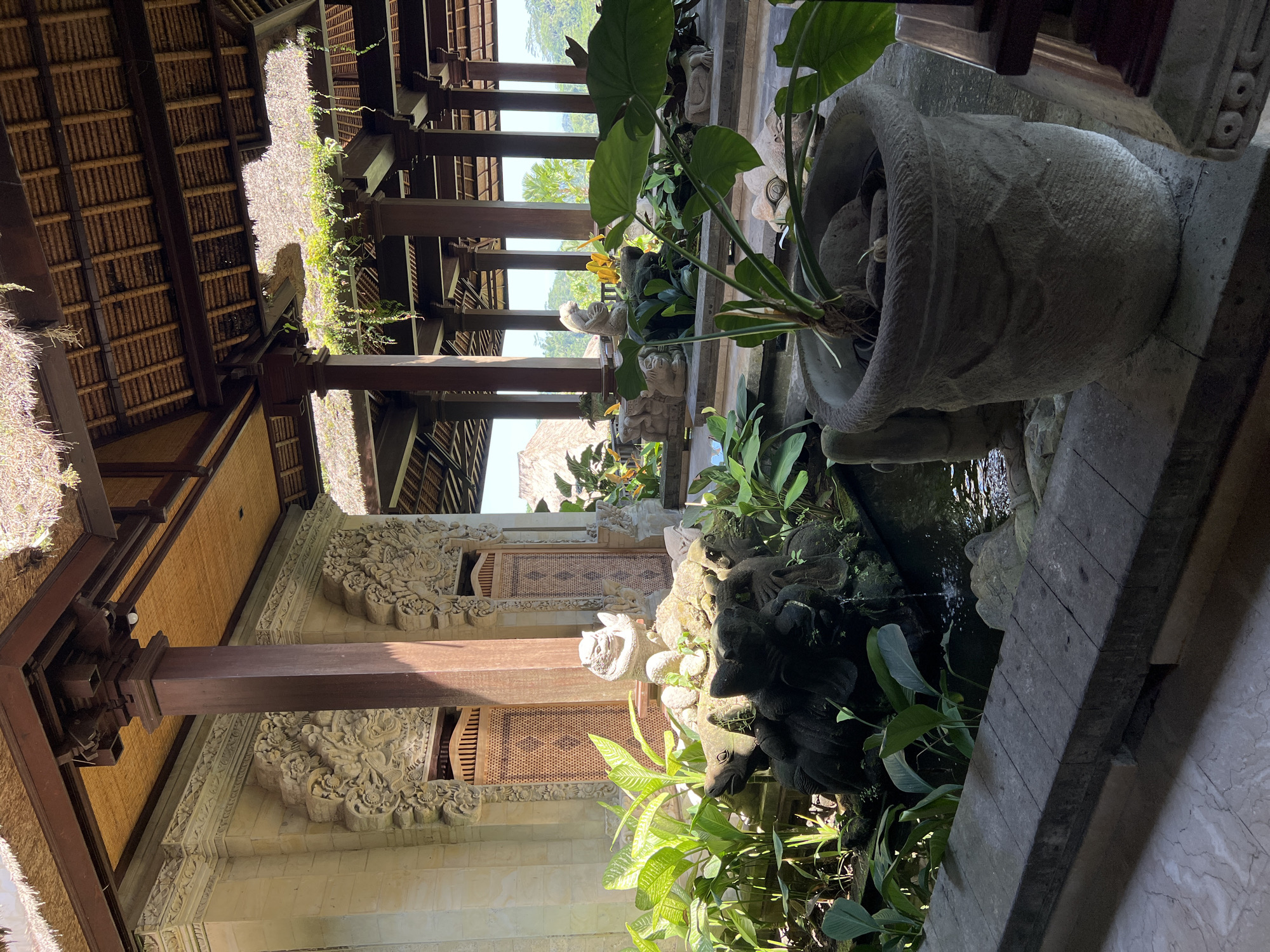 The image depicts an outdoor space in Kedewatan, Indonesia. It features traditional Balinese architecture, including ornate stone carvings and a thatched roof. A water feature, adorned with statues and lush vegetation, is prominent. A large stone planter with greenery is positioned near the edge of the water feature. The architecture includes wooden beams.