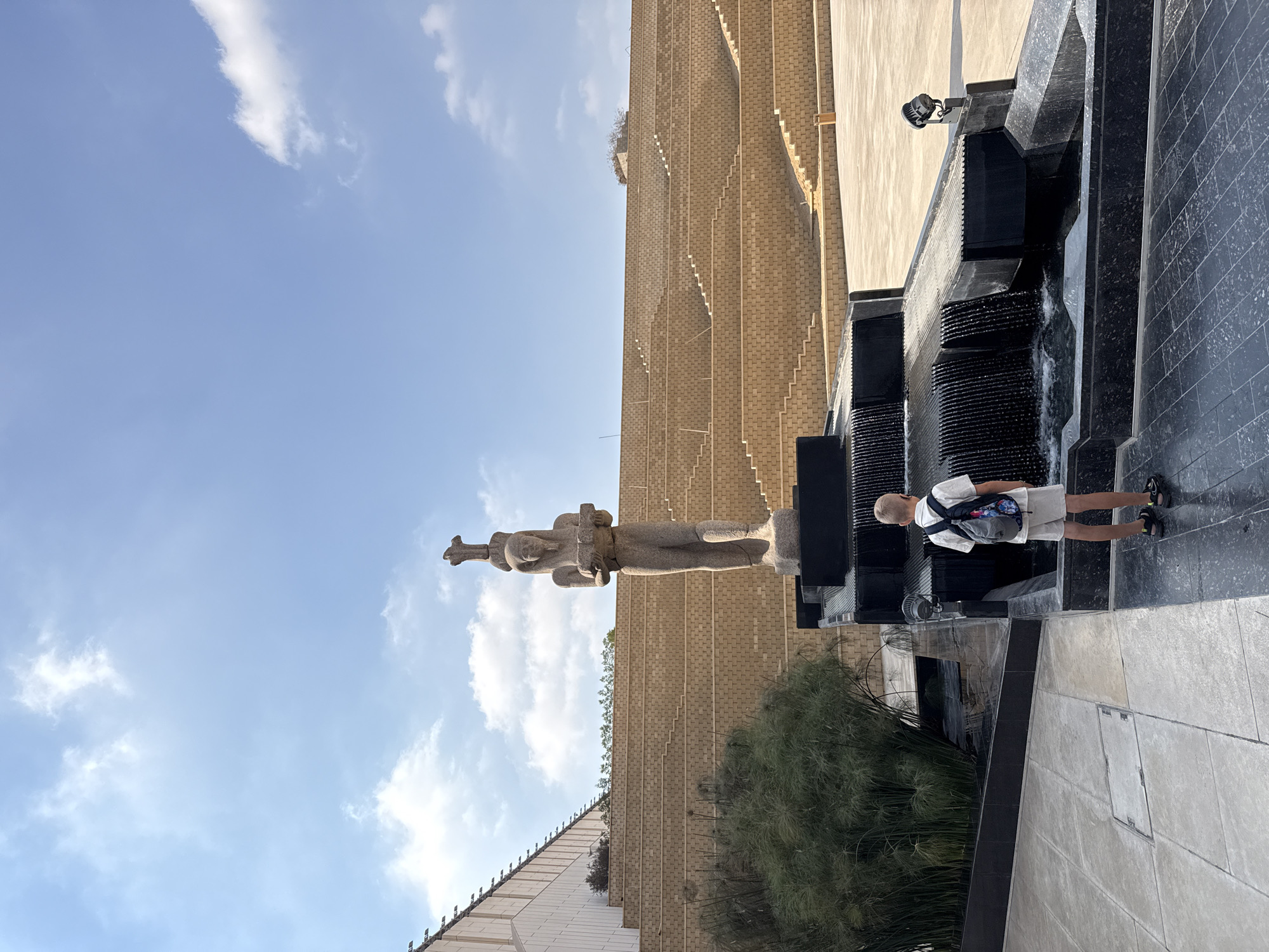 A young boy with a backpack stands with his back to the camera, facing a multi-tiered fountain with cascading water. In the background, a large statue of an ancient Egyptian figure stands before tiered seating made of light brown brick. The scene is set in an open, paved area under a bright blue sky with scattered clouds. The location is identified as Kafrat Nassar, Egypt.