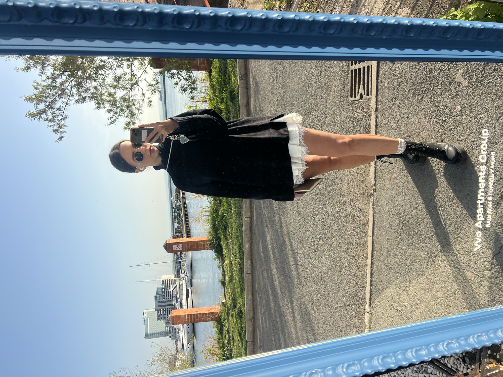 A woman is seen in a mirror reflection. She is holding a phone and wearing sunglasses, a black top, a white skirt, and black boots with white socks. The mirror is framed in blue and is reflecting an outdoor scene. The backdrop features a cityscape, bodies of water with boats, and green vegetation. The area in the foreground appears to be a paved surface. A building with Cyrillic text "VVO Apartments Group" is partially visible, suggesting the location is Vladivostok, Russia.