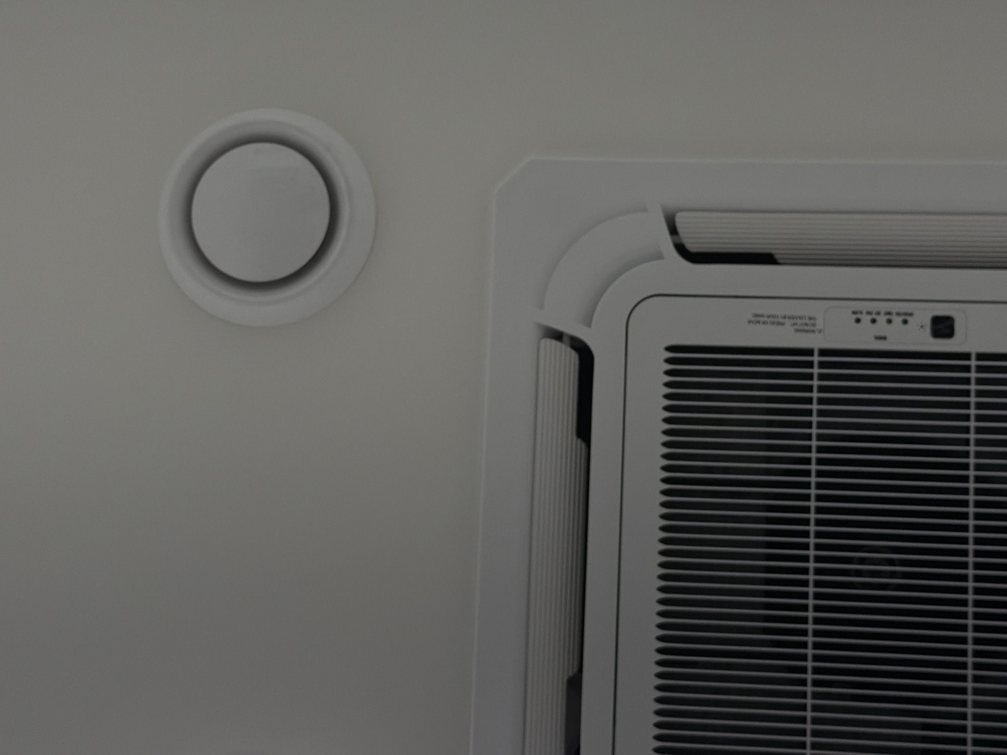 The image shows a partial view of a ceiling with an air vent and an air conditioning unit. The air vent is circular and white, situated on the left side of the image. To the right is a white air conditioning unit, exhibiting a grill pattern and a control panel with indicator lights. The scene is devoid of people and lacks contextual elements indicating a specific location like Izhevsk, Russia.