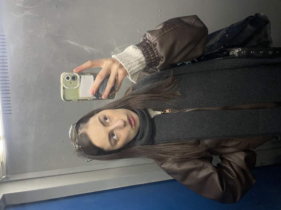 A female individual is depicted taking a mirror selfie within what appears to be an elevator or small utilitarian room. Her dark brown hair is parted centrally with sunglasses resting on her head. She wears a brown leather-style bomber jacket, a light-colored ribbed sweater with elasticated cuffs, and a dark grey ribbed scarf. A gold-zippered garment is visible beneath the scarf. She holds a smartphone with a light green case, featuring "GOOD LIFE" text, a landscape image, and a white sticker. Her gaze is directed upwards and to her left, away from the camera. A black bag with a zipper and eyelets is slung over her left shoulder. The background consists of a metallic-grey reflective surface, a vertical blue panel or door frame on the left, and a horizontal metallic vent above. The specific location within Moscow, Russia, cannot be determined from the visual information provided.