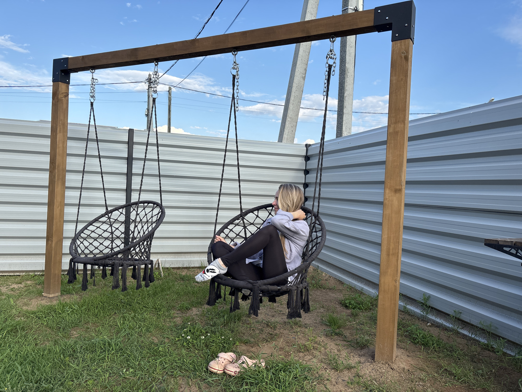 A young woman with blonde hair is sitting in a black macrame hanging chair in an outdoor setting. She is wearing a light blue long-sleeved shirt, black leggings, and gray socks with a Mickey Mouse design. The chair is suspended from a wooden frame with metal brackets. To the left of her chair, another similar black hanging chair is visible. The ground is covered in green grass and dirt. In the background, there is a corrugated metal fence and tall utility poles with power lines. The sky is blue with some white clouds. A pair of pink sandals are on the ground near the woman's chair. The scene appears to be a backyard or a recreational area, possibly in the city of Chaikovsky, Russia, given the context.