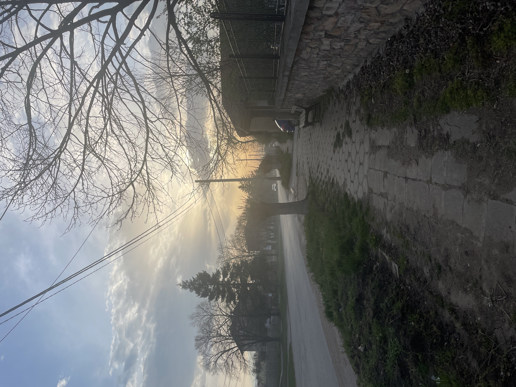 The image shows a street scene in Aprilovo, Bulgaria. The view is along a sidewalk and street, looking into the distance. Bare tree branches are visible in the upper left corner and throughout the center of the frame. Above, there is a sky with some clouds and fog, the sun is obscured by the fog. The sidewalk, made of hexagonal paving stones, runs along the right side of the image next to a stone retaining wall with a metal fence. On the other side of the sidewalk is a grass and dirt area. The street, which appears to be asphalt, runs parallel to the sidewalk. In the distance, there are more houses and trees. Power lines cross the upper center of the image.