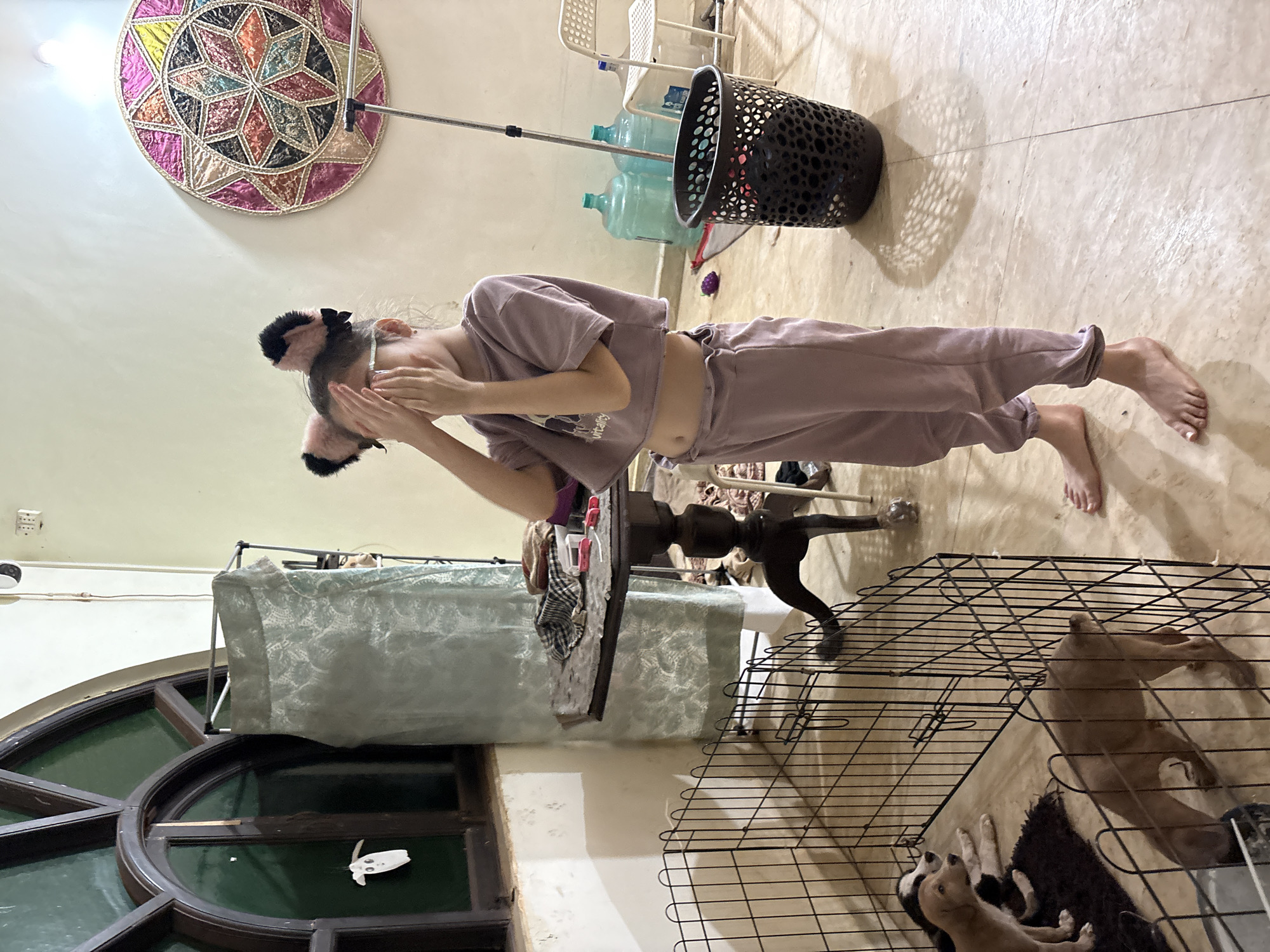 A young girl wearing a pink crop top and matching sweatpants, adorned with fluffy cat ears, stands in what appears to be a covered patio area. She is covering her eyes with her hands, suggesting she is playing a game or hiding. To her left, a black wire puppy pen contains three puppies, one of which is a light brown color, another is brown and white, and the third is dark brown. In the background, several large water jugs, a black trash can, and a white chair are visible. A round, colorful woven tapestry hangs on the wall. The floor is tiled, and a wooden table with various items on it, including a checkered cloth and some pink objects, is positioned behind the pen. The overall environment suggests a domestic setting in Ashvem, India.