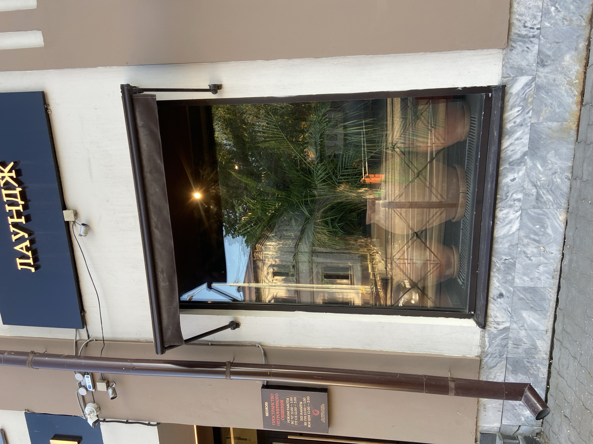 The image depicts a window of the NEGRONI establishment in Kazan, Russia. The window reflects a building across the street and interior elements, including a palm tree and large clay pots. A dark awning partially covers the window. Below the window, there's a sign in both Russian and Tatar languages stating the name of the place, NEGRONI, "a space for Negroni communication", and its operating hours. A brown drainpipe runs alongside the building's facade, and security cameras are visible above the pipe. Marble pavement surrounds the base of the building. A sign that says “ЛАУНДЖ” is also visible.