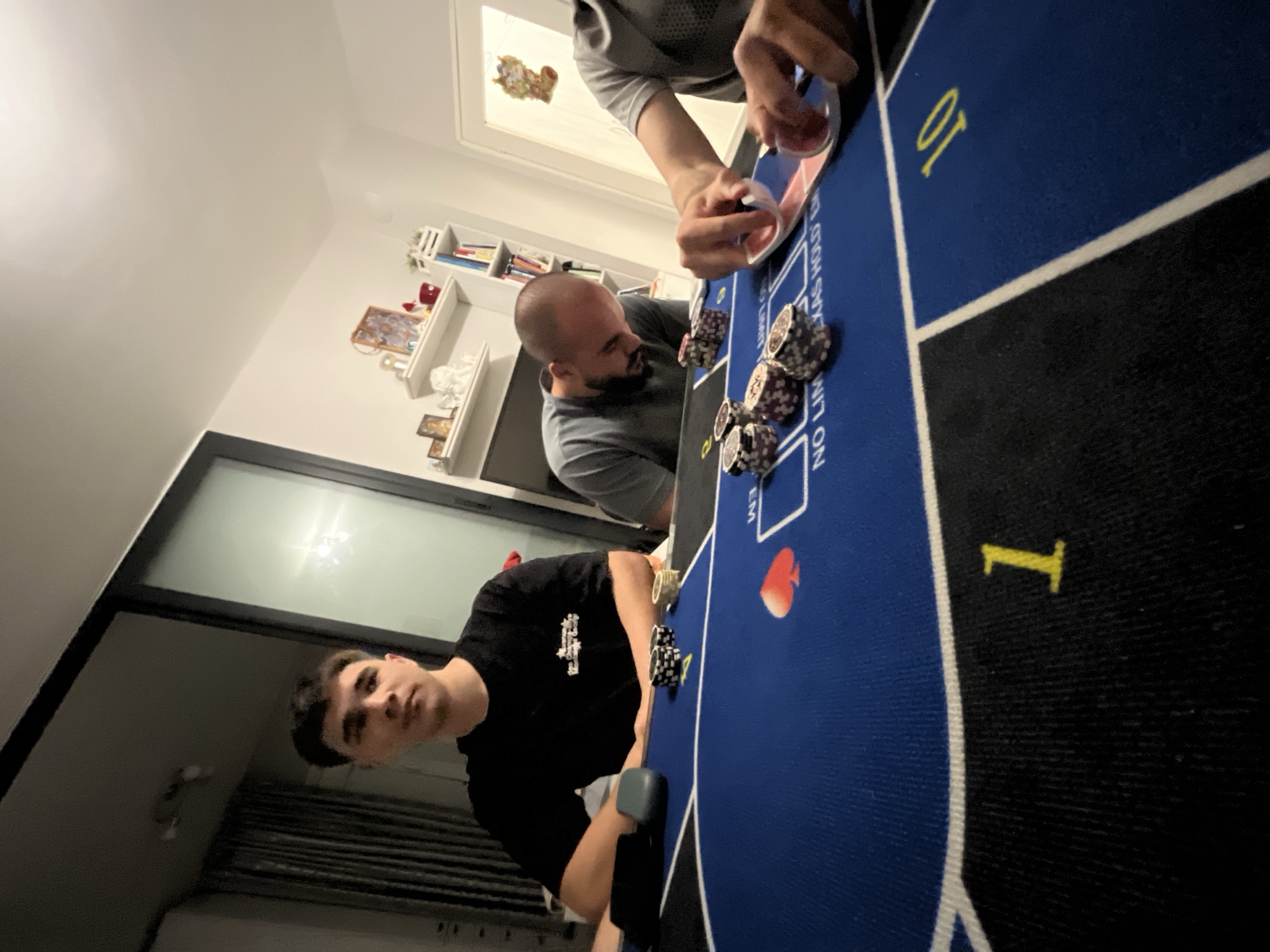 Here is a detailed description of the image:
**Scene:**
The image captures a poker game in progress. Three individuals are visible around a blue poker table. The table has markings including "NO LIMIT TEXAS HOLD EM" and numerical indicators (10, 1). Stacks of poker chips are scattered around the table.
**People:**
* **Individual 1:** Located in the foreground, wearing a black t-shirt.
* **Individual 2:** Partially visible, seated at the side of the table, wearing a gray t-shirt. Bald.
* **Individual 3:** Top of head and hands visible.
**Actions/Interactions:**
* Individuals are engaged in a poker game. One is holding cards.
**Environment:**
The game appears to be taking place indoors, possibly in a home. A window is visible in the background. There is a bookshelf/cabinet with decorative items.
**Location:**
There is not information available in the picture to ascertain its location, or the location of Burgas, Bulgaria.