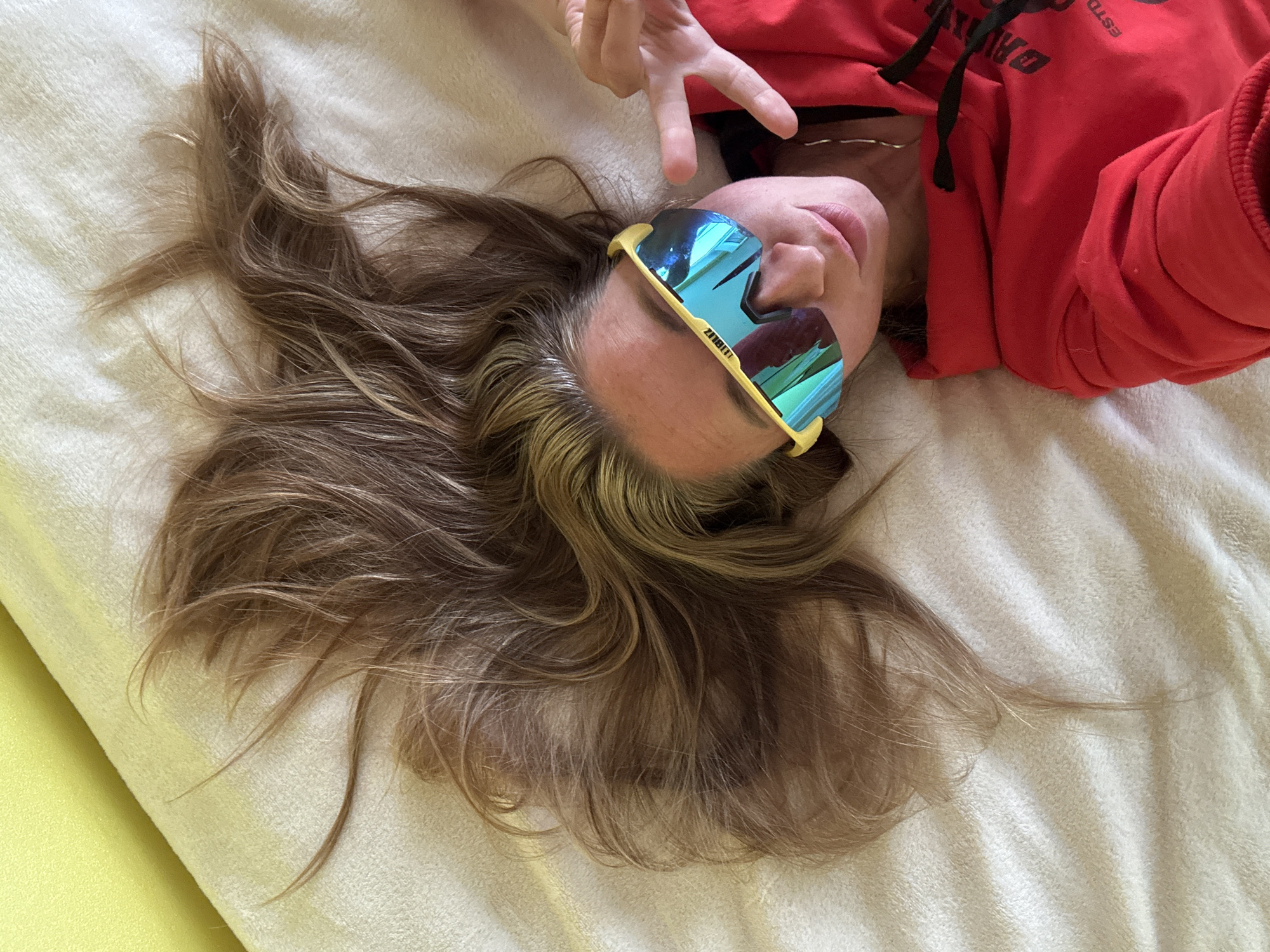 A person is lying on a soft, textured, cream-colored surface, possibly a blanket or bed. Their head is tilted back, and their long, wavy, light brown hair is spread out around them. They are wearing a bright red hooded sweatshirt. On their face, they are wearing bright yellow-rimmed sunglasses with reflective blue lenses. Their right hand is raised, with two fingers extended in a peace sign. The person's mouth is slightly open. The lighting suggests it could be daytime indoors, with soft shadows indicating indirect light. There is no visible indication of the weather or specific location cues, other than the provided context of Прикамский, Russia. The overall mood appears relaxed and playful. The sunglasses have text visible on the frame, which appears to be "BLIZ". The red sweatshirt has some dark text on it, but it is partially obscured and difficult to read clearly.