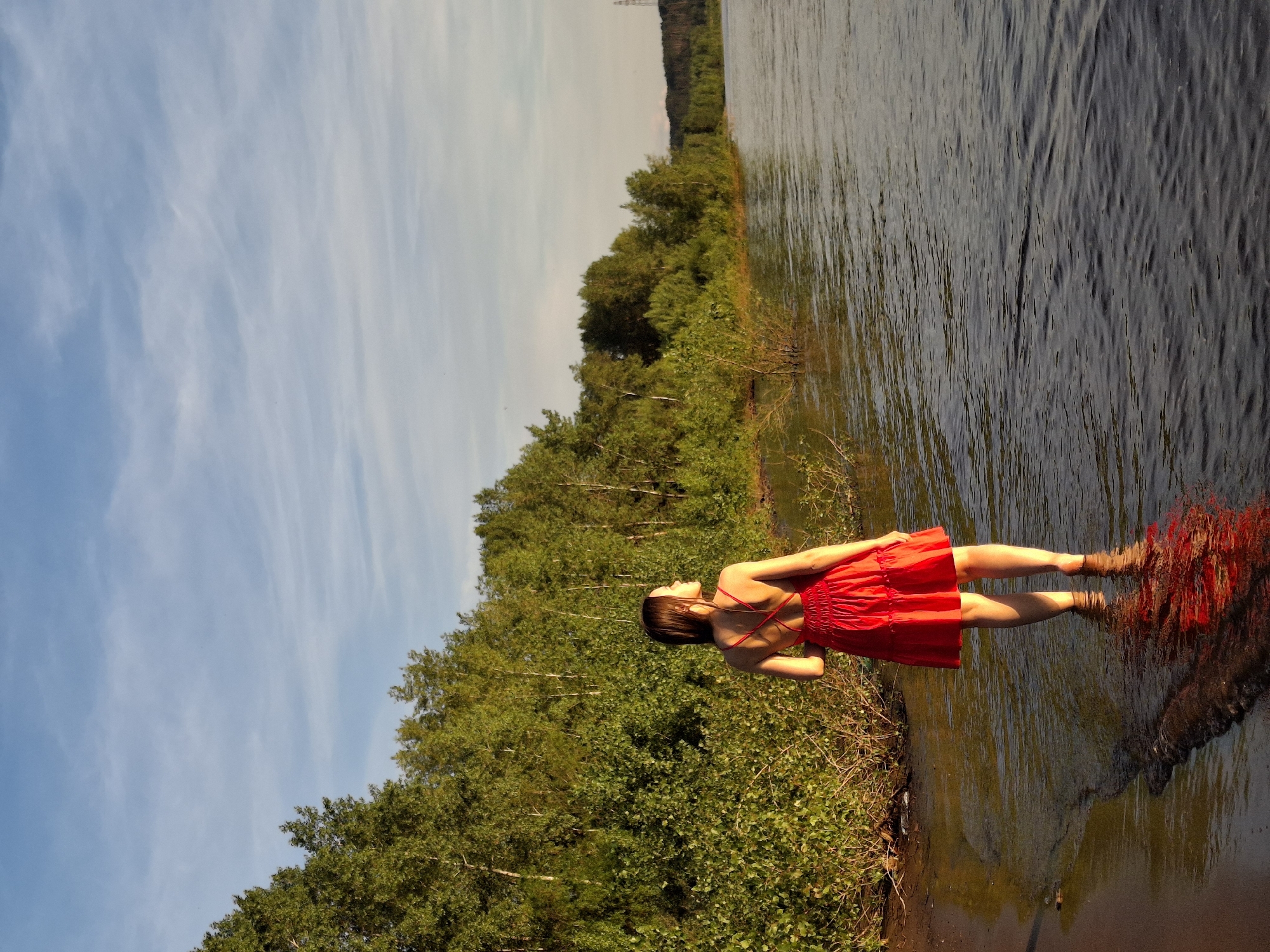 In the city of Chaikovsky, Russia, a young woman stands in shallow water near the shore of the Kama River. She is wearing a bright red dress with a low back and her feet are submerged in the water. The riverbank is lined with lush green trees and vegetation. The sky above is a clear blue with scattered white clouds, illuminated by the warm glow of the setting sun. The reflection of the woman and the surrounding greenery is visible in the calm water. The scene captures a peaceful moment by the river at dusk.