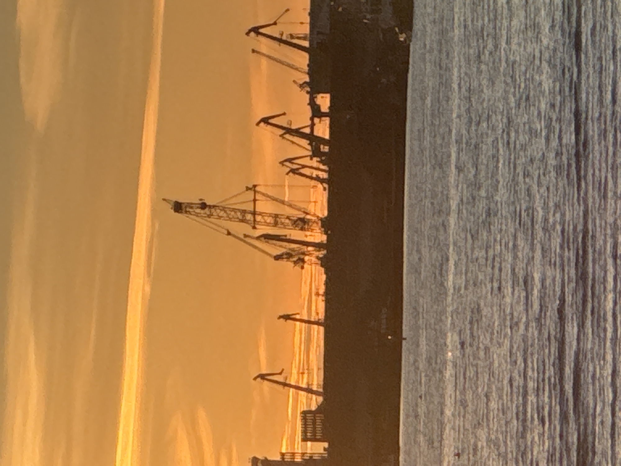 The image depicts a harbor scene at sunset in Tallinn, Estonia. The dominant subjects are several large cranes silhouetted against a warm, orange and yellow sky. The sky is filled with soft, elongated clouds that are illuminated by the setting sun, creating a dramatic and serene atmosphere.
In the foreground and to the right, the calm waters of the harbor are visible, with the textured surface reflecting the ambient light and creating vertical lines. The juxtaposition of the industrial cranes and the natural beauty of the sunset suggests a functioning port during a peaceful time of day.
The silhouette of a dock or pier is also visible, with some structures and railings partially discernible. The overall impression is one of quiet industrial activity at the end of the day, bathed in the golden light of sunset. There is no visible text in the image.