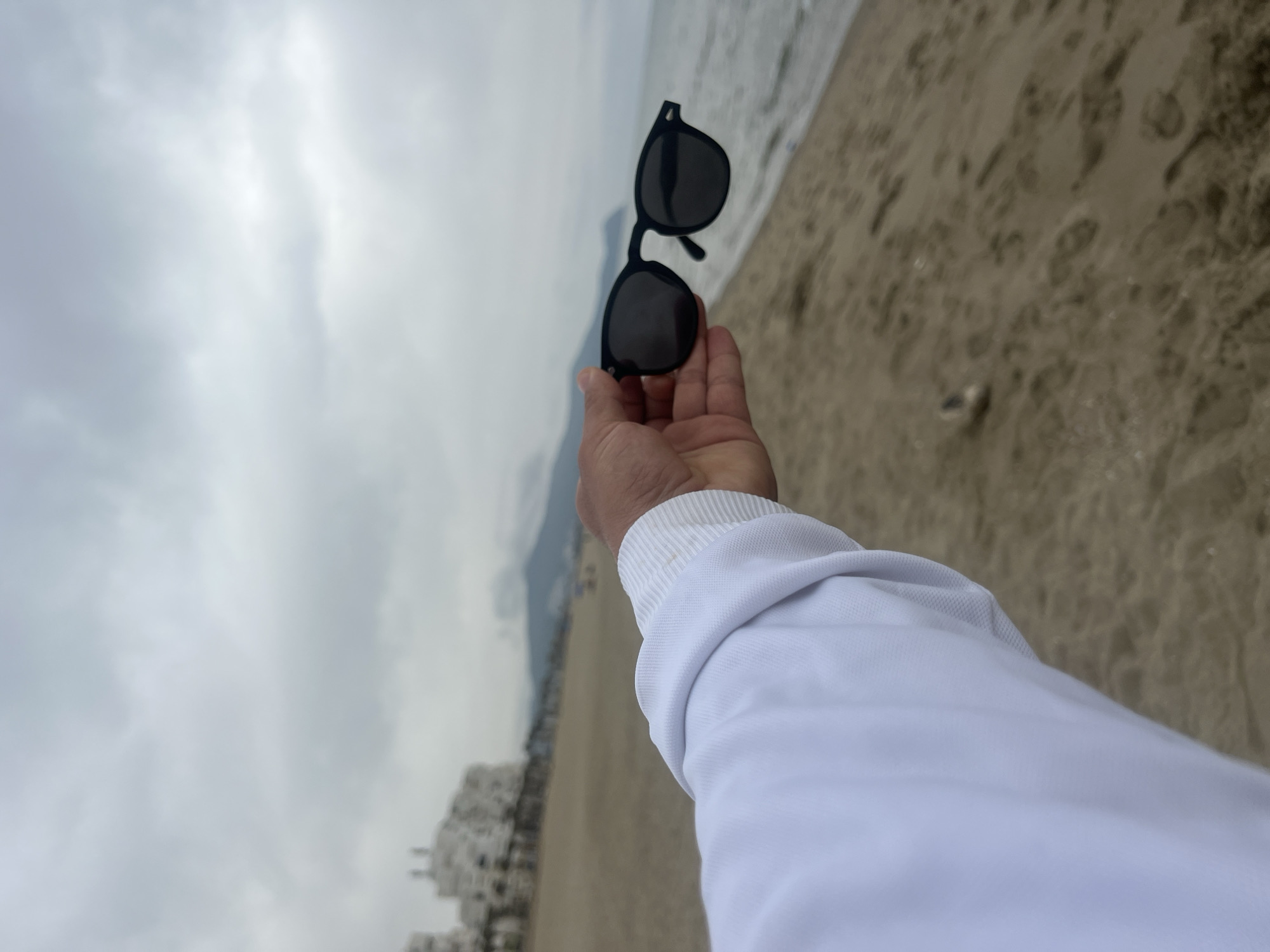 The image, viewed from a first-person perspective, captures a person's right hand holding a pair of black sunglasses upwards. The hand and lower forearm, covered by a white long-sleeved shirt or sweatshirt with a ribbed cuff, dominate the foreground. The sunglasses have dark, possibly polarized, lenses and appear to be a cliplike design.
The background reveals a wide, expansive beach scene under an overcast, cloudy sky. To the right, sandy shore meets the calm waters of the ocean, with a gentle waterline visible. In the mid-ground, a line of white buildings, possibly part of a coastal town or resort, stretches along the beach. Distant mountains are faintly visible beneath the heavy cloud cover. The diffused light suggests a cloudy day rather than bright sunshine. The setting is consistent with a coastal area like Martil, Morocco. There is no visible text in the image.