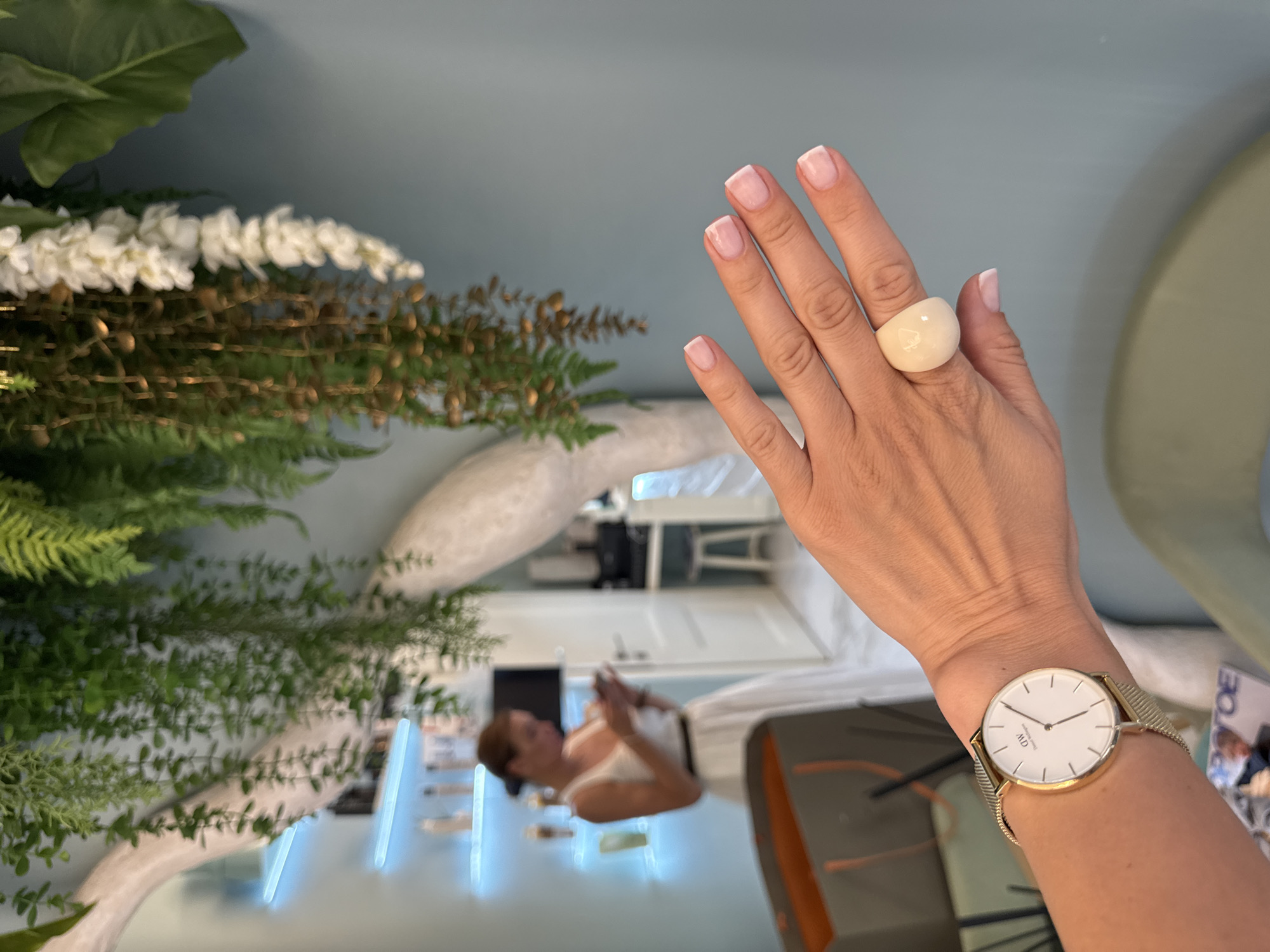 The image depicts a close-up of a woman's hand wearing a gold watch and a large, rounded, pale ring. The hand has manicured nails with a French tip. In the background, a woman in a white dress is visible, looking at her phone. The environment appears to be an indoor space with shelves of products, a decorative tree-like structure, and hanging plants. The wall color is a muted blue. There is also a glimpse of a shopping bag and a magazine on a surface. The overall impression is that of a salon or spa. The scene is set in Sochi, Russia, although no specific landmarks are identifiable from the image.
