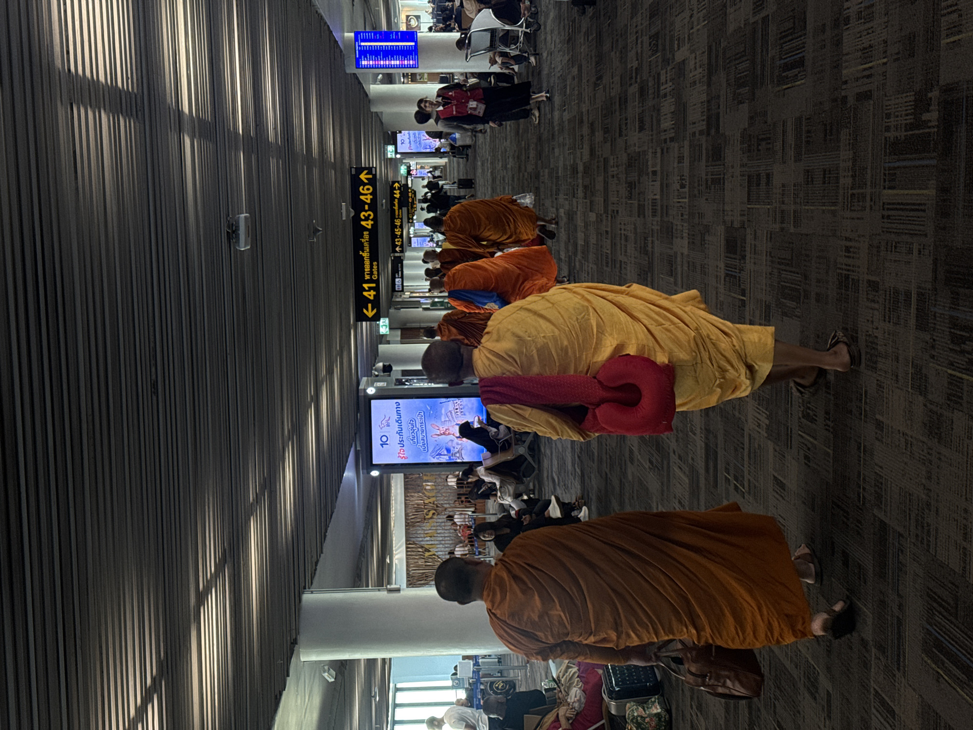 This image captures a moment at what appears to be a busy airport terminal, likely in Bangkok, Thailand, given the Thai text visible. The scene is dominated by several Buddhist monks in saffron and orange robes, sitting or standing in the midst of other travelers. The monks' presence suggests a religious or cultural aspect to their journey, perhaps a pilgrimage or travel related to their monastic duties.
The setting is an indoor airport concourse with a distinctive patterned carpet. Overhead, a modern ceiling with vertical ribbed panels casts long shadows, indicating a time of day when sunlight is angled, perhaps late afternoon or early morning. Large digital signs display flight information and other announcements, one of which has Thai text and the number "10". Another sign in Thai and English indicates gate numbers "41" and "43-46".
The overall mood is one of quiet contemplation amidst the general hustle and bustle of air travel. Some monks are seated, appearing relaxed, while others may be waiting for their flights. The other travelers are seen in the background, some with luggage, engaged in typical airport activities like waiting or walking. The image is well-lit, allowing for clear visibility of the subjects and their surroundings.