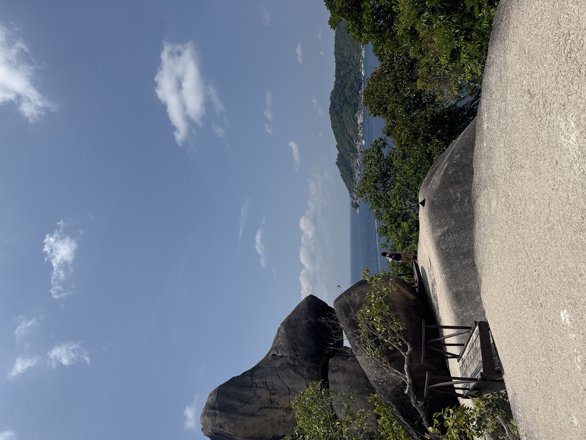 A photograph depicts a scenic overlook in Lam Kaen, Thailand. It features large granite boulders in the foreground, partially covered with greenery. A small wooden observation platform is affixed to one of the boulders. A person stands on the edge of the boulder looking out towards the sea and an island in the distance. The sky is blue with scattered clouds. The photograph appears to be taken from a low angle, emphasizing the height of the rocks.