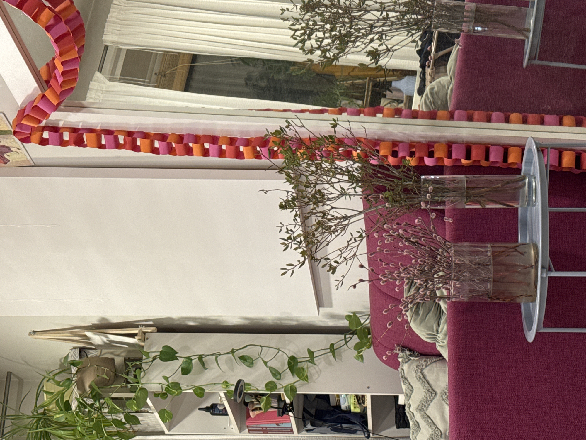 The image depicts an interior scene, presumably in a home. Dominating the view is a white wall on the left side of the image, and in front of it, there is a set of shelves with plants and other household objects on it. A red couch with pillows on it is on the right side of the image. There are vases with branches in them on the right side of the image, on top of a silver tray. There is a pink and orange paper chain hanging near the top of the image. It is not possible to confirm the image was taken in Krasnoyarsk, Russia.
