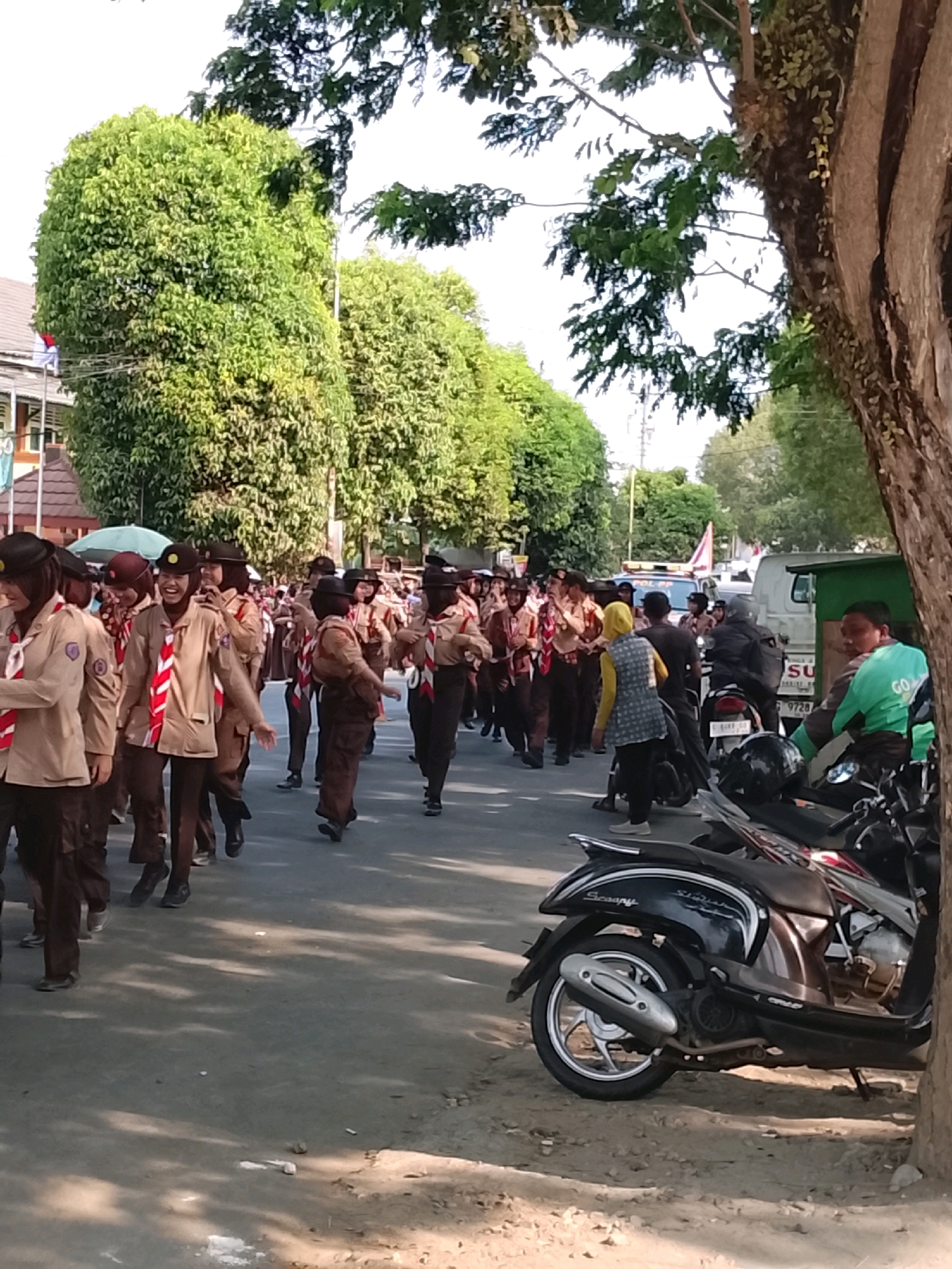 The image depicts a street scene in Slawi, Indonesia, with a group of people, likely students in scout uniforms, participating in a parade or procession. The individuals are wearing brown uniforms with red and white neckerchiefs and black hats. They are walking along a paved road lined with trees and buildings. Several motorcycles are parked along the side of the road, and a few people are observing the procession from the roadside. The sky is visible in the background, suggesting it is daytime. The overall atmosphere appears to be one of a community event.