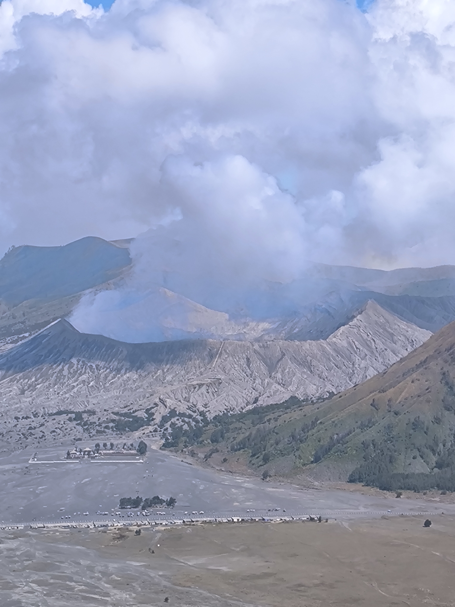 This is a medium shot of a volcanic landscape under a cloudy sky. In the foreground, a wide, flat expanse of gray sand and dirt stretches out, dotted with small groups of people and parked vehicles. Several long, low structures, possibly tents or temporary shelters, are set up in neat rows along the edge of this expanse. Further back, a collection of small buildings, some with pointed roofs, are clustered together amidst a few scattered trees.
Beyond this, the terrain rises dramatically into rugged, barren mountains. The slopes are a light gray, textured with numerous ridges and gullies, suggesting a history of volcanic activity. At the center of the frame, a cone-shaped volcano emits a thick plume of white and gray smoke that drifts upwards and to the left, partially obscuring the view of the surrounding peaks. The sky above is filled with large, puffy white and gray clouds, with hints of blue sky peeking through in places. The lighting suggests it is daytime, possibly morning or late afternoon, with the sun diffused by the cloud cover. The overall mood is one of natural grandeur and the raw power of the earth, with a hint of human presence in the organized settlement in the foreground. No visible text is present in the image.