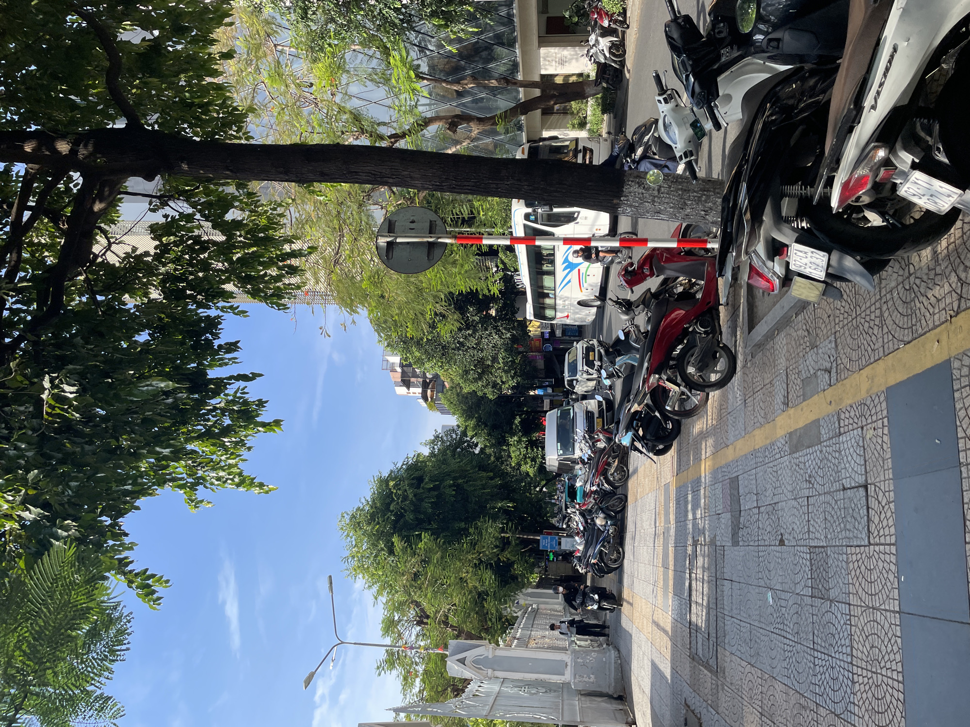 The image depicts a street scene in Da Nang, Vietnam. In the foreground, a sidewalk paved with patterned tiles is visible, with several parked motorcycles. A yellow line runs along the edge of the sidewalk. Further back, a white bus is driving on the street, alongside other vehicles and more parked motorcycles. Lush green trees line the street, with a building featuring a modern glass facade visible behind the trees. The sky is blue with scattered clouds. Two individuals are seen walking on the sidewalk near a white decorative gate. One person is riding a black motorcycle. The overall scene suggests a typical urban environment in a Vietnamese city on a sunny day.