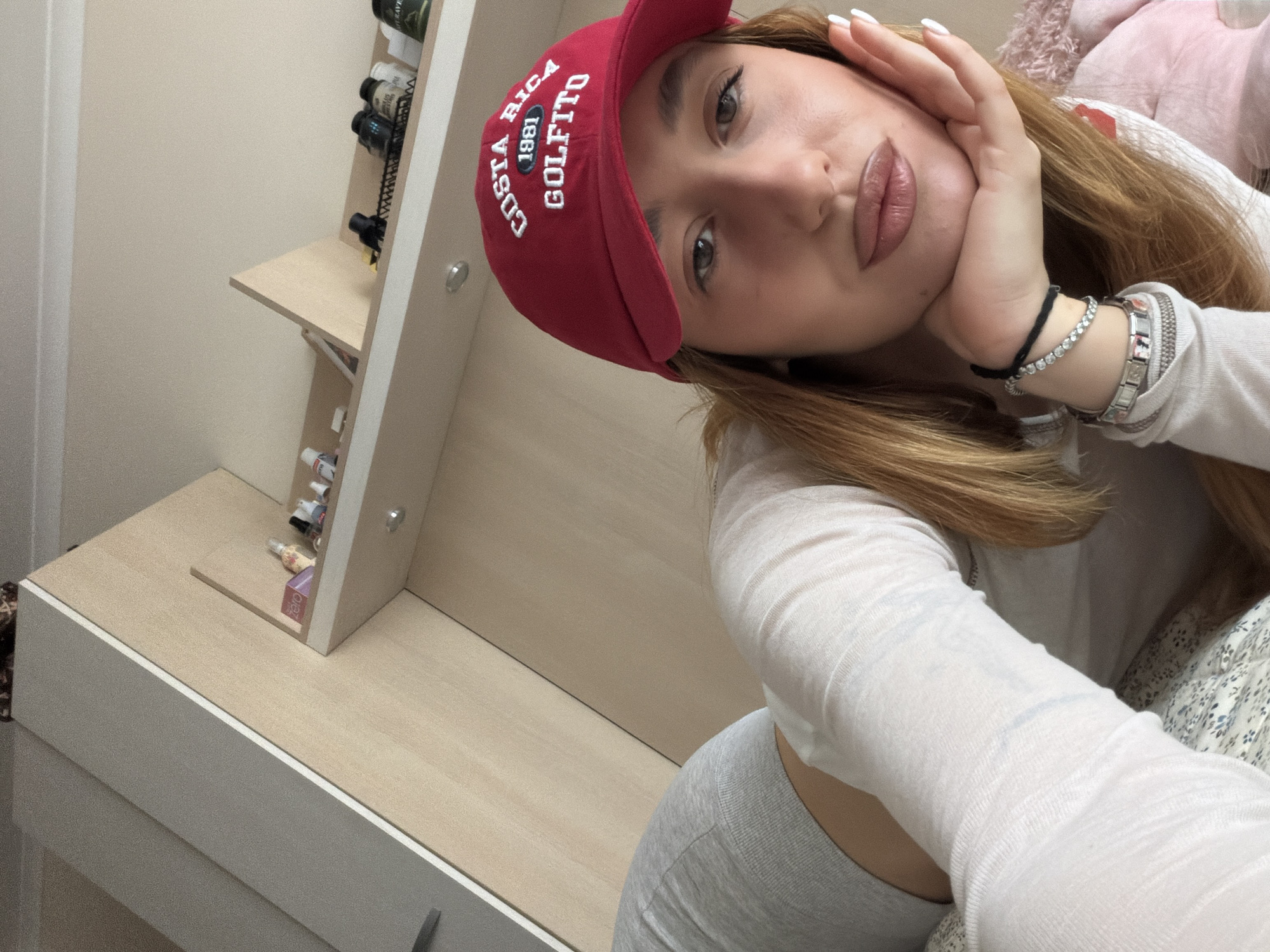 A young woman wearing a red cap with "COSTA RICA 1981 GOLFITO" embroidered on it poses for a selfie. She has fair skin, light brown hair, and is wearing a white long-sleeved shirt and gray leggings. She is wearing bracelets on her left wrist. Her face is tilted, and she is resting her head on her hand. The background includes a white cabinet with shelves holding small bottles and jars. There is also a partially visible bed with a floral patterned blanket and a pink stuffed animal. The image does not provide any information about the location being Sofia, Bulgaria.